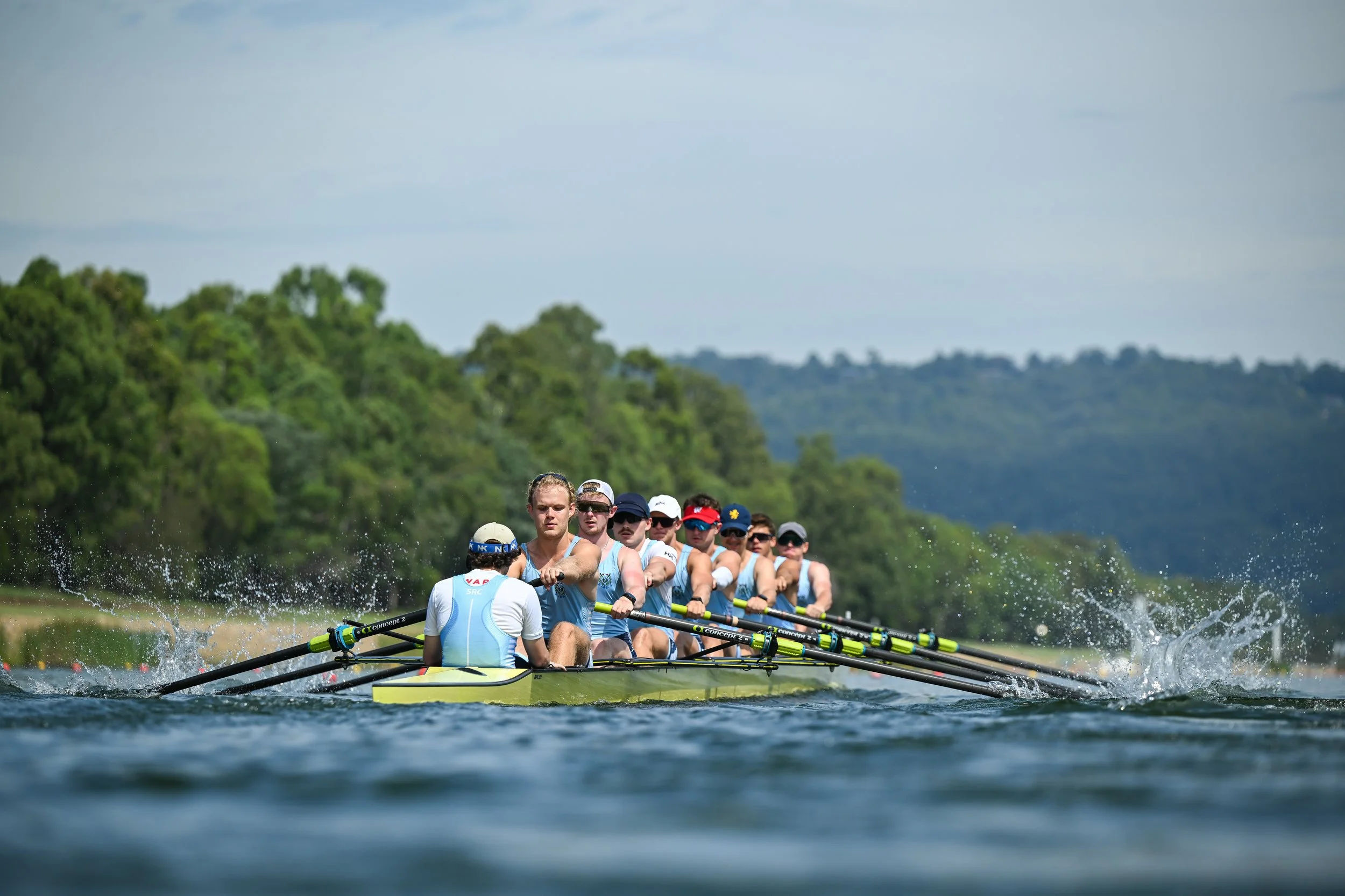 A rowing team practicing on a river surrounded by green trees, with several rowers in a yellow boat pulling oars in sync.