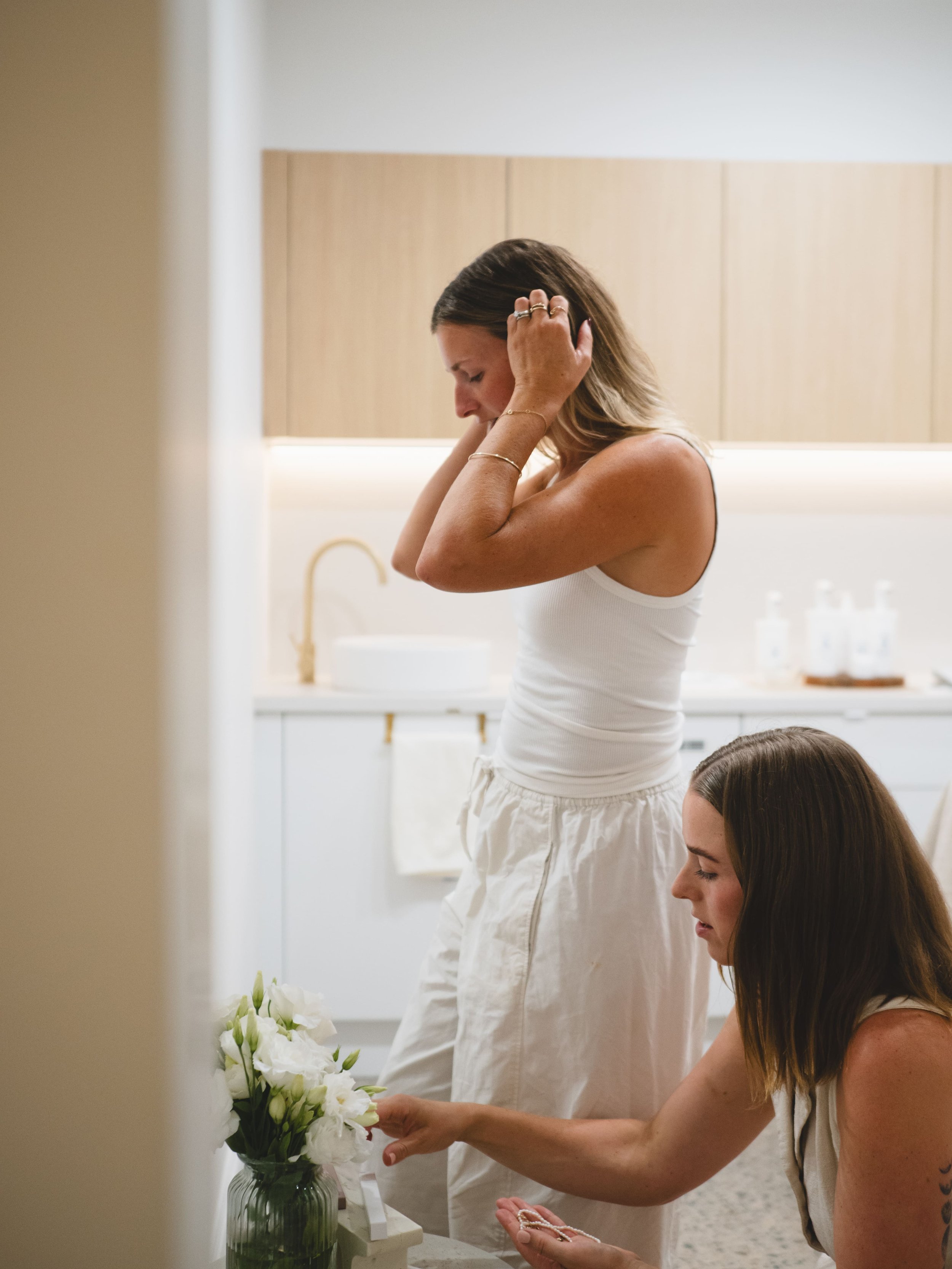 Two women in a minimalist kitchen; one is fixing her hair and the other is arranging white flowers in a vase.