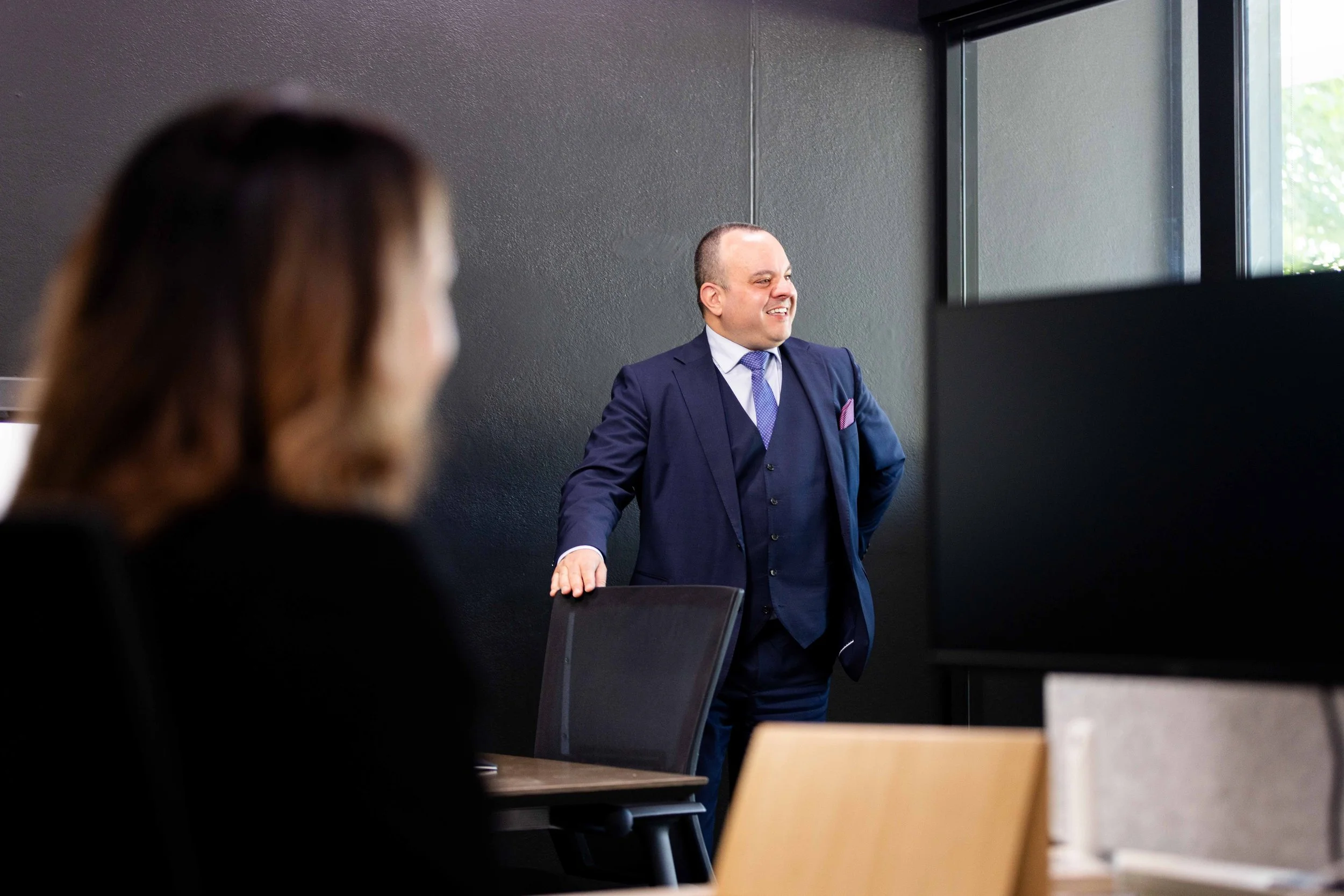 Man in a blue suit presenting or speaking in a meeting room with two colleagues, one partially visible, and a window in the background.
