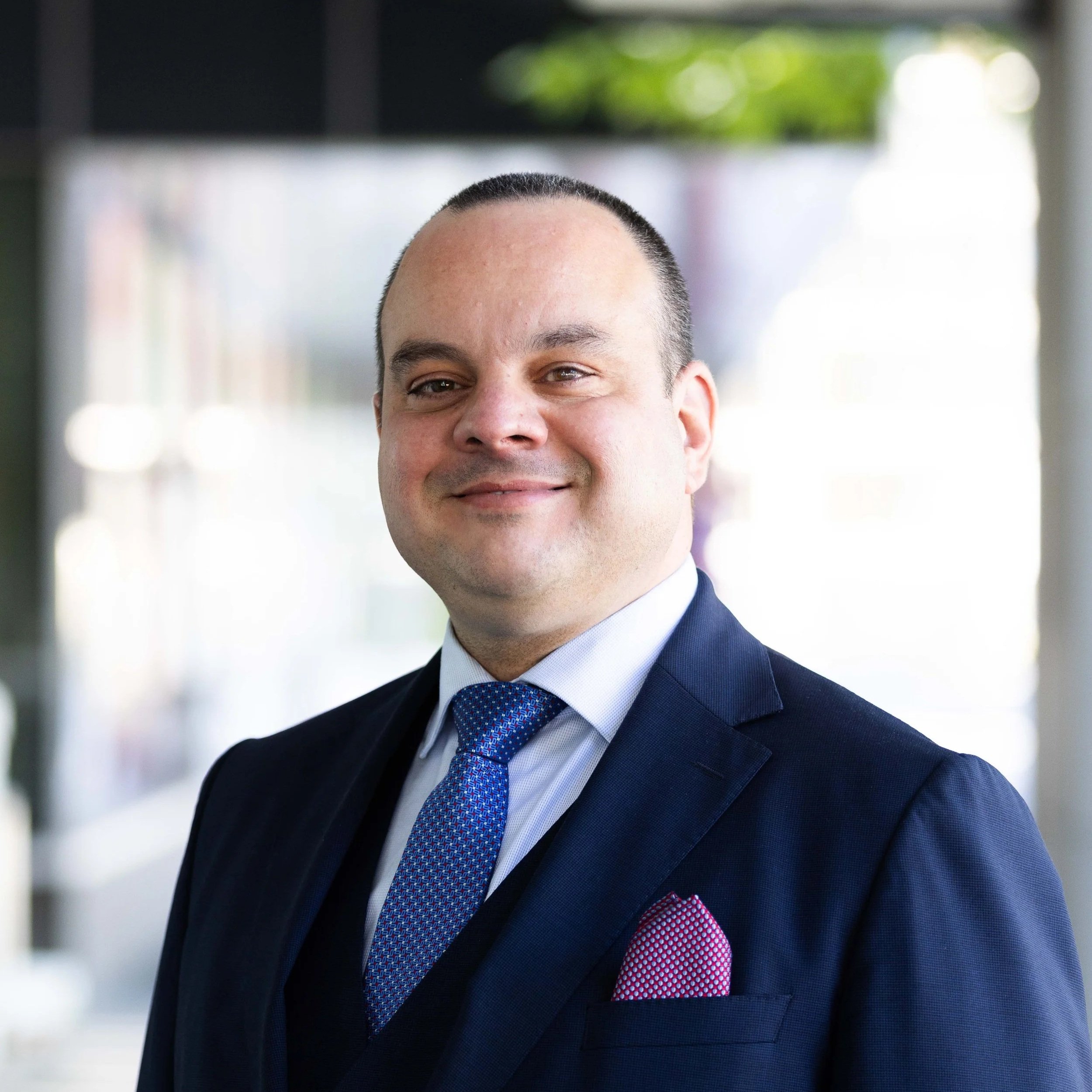 A man wearing a navy suit, light blue dress shirt, and a blue patterned tie, smiling outdoors with a blurred background of trees and buildings.