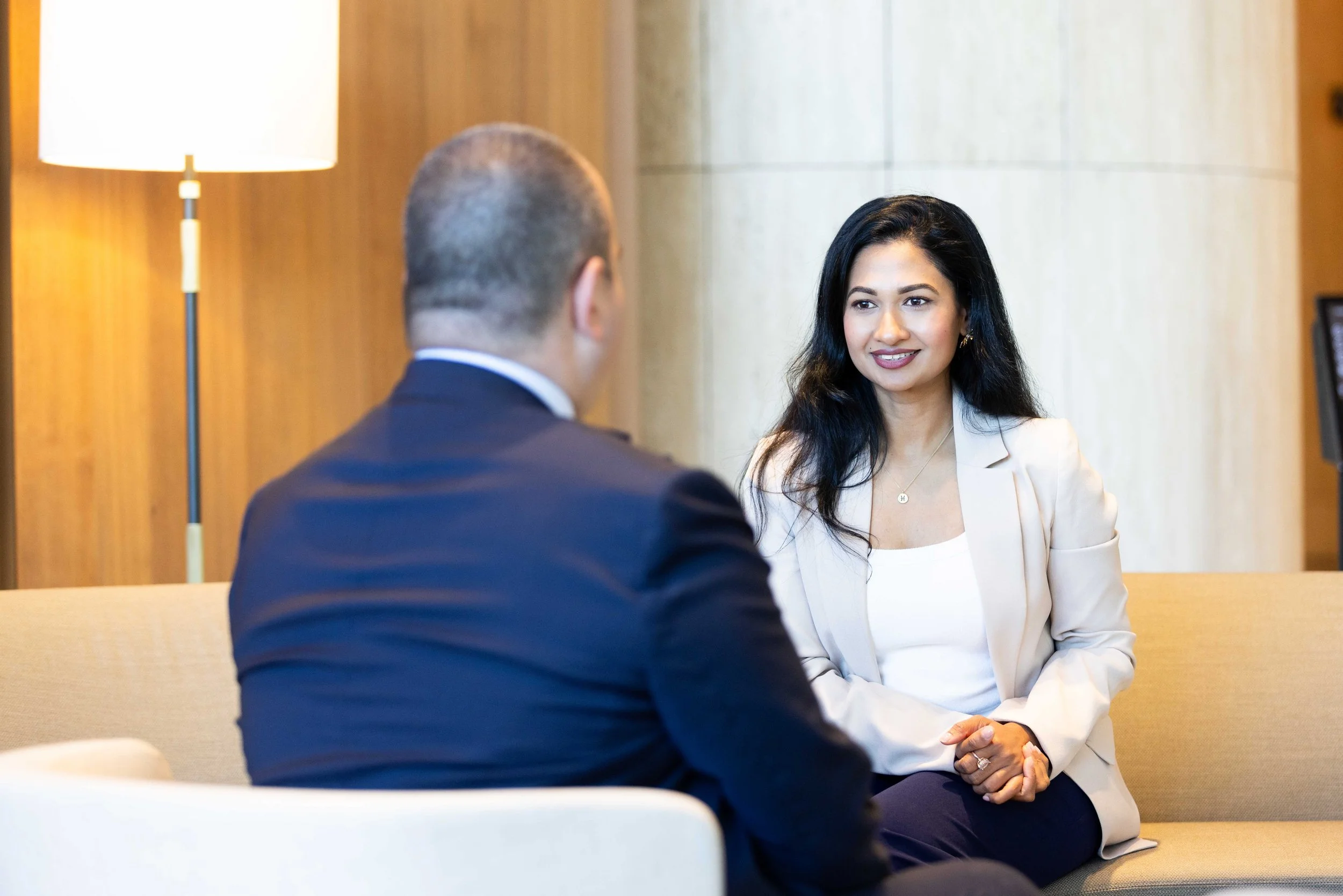 A woman with black hair wearing a cream blazer and white top talking to a man in a dark suit, sitting on a beige sofa in a room with wooden and cream walls, with a floor lamp in the background.