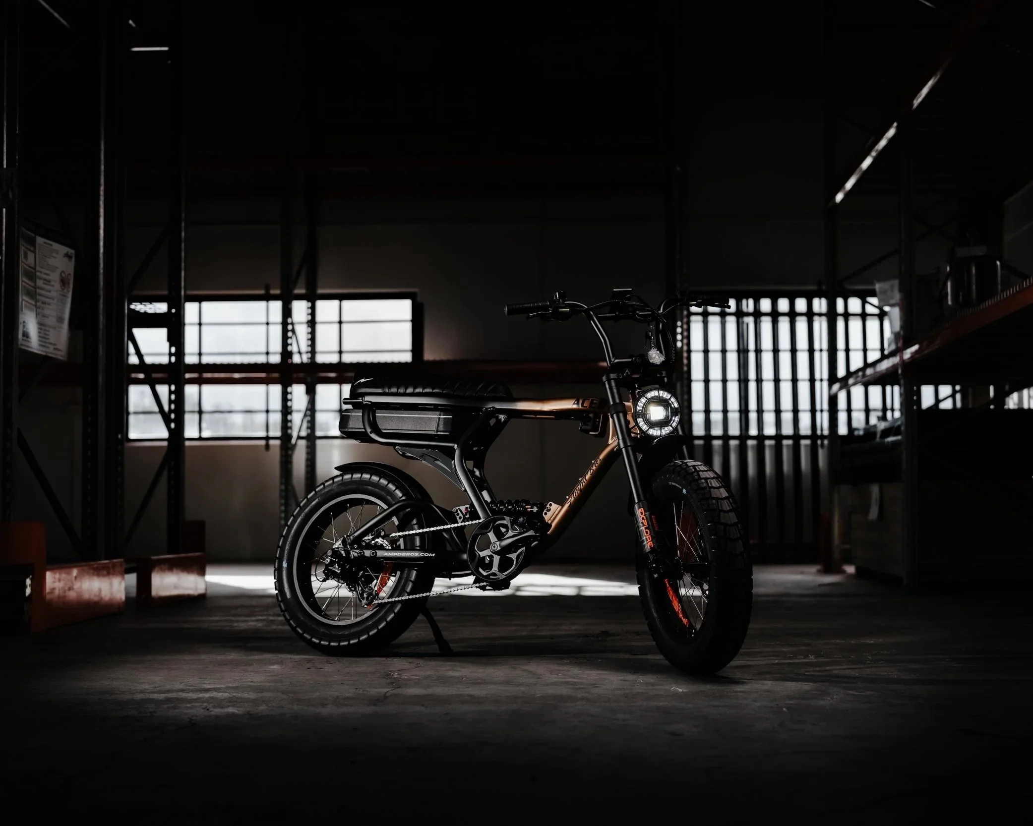 Electric bicycle with headlights on, placed in a dark warehouse with large windows, metal shelving units, and orange crates.
