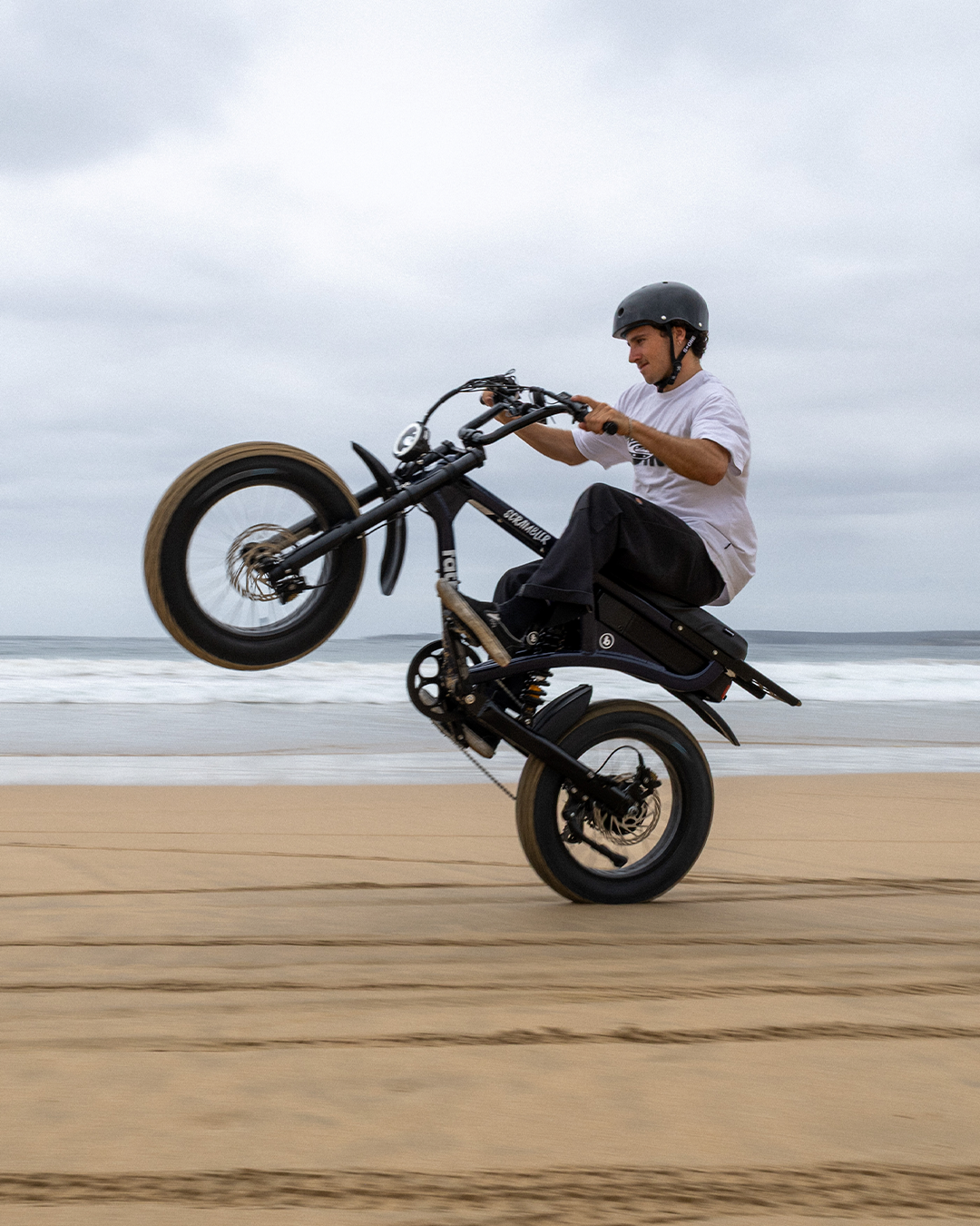 A man riding an electric bike on a beach, performing a wheelie with his front wheel lifted off the ground. The sky is cloudy, and the ocean is visible in the background.