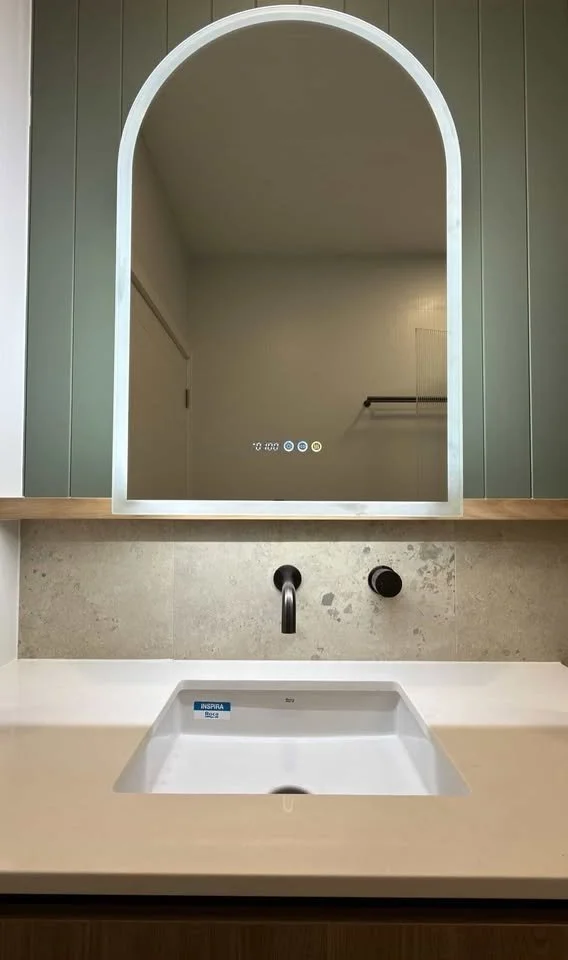 Modern bathroom vanity with a white rectangular sink, black faucet, backed by gray textured tile, with a large arched mirror above.