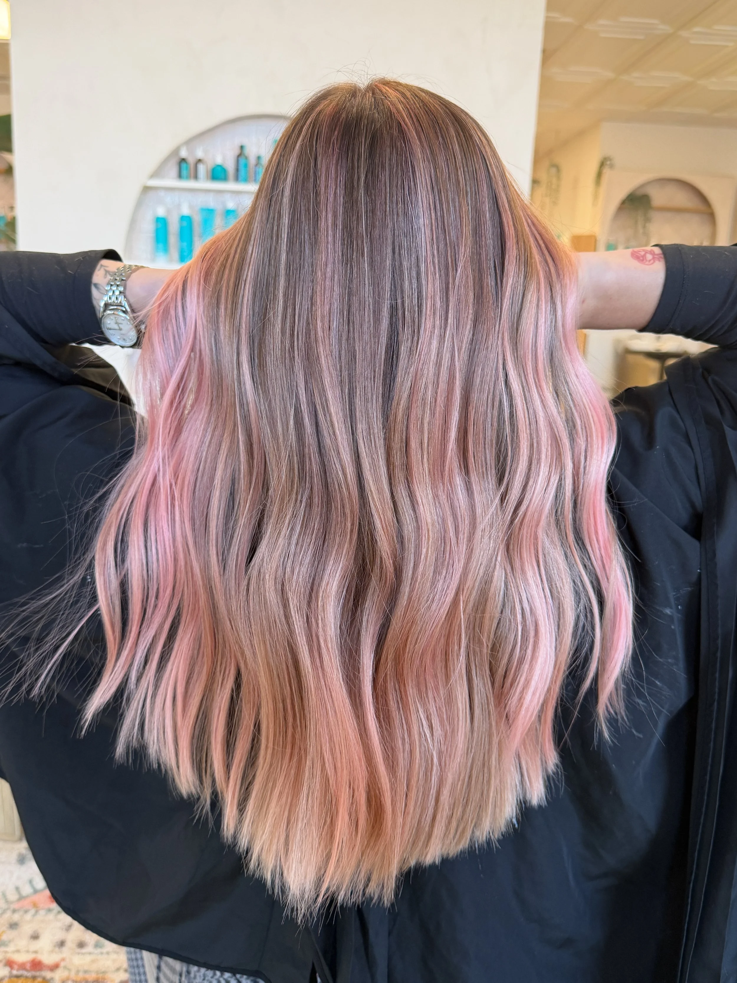 A woman with long wavy hair that is pink and blonde, with a darker root, seen from behind in a salon.