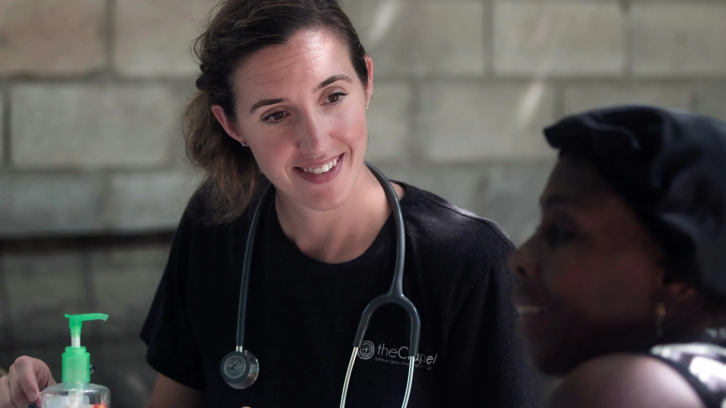 A woman healthcare professional talking to a patient, both smiling, in a clinical setting. The healthcare worker is wearing a black shirt with a logo and has a stethoscope around her neck.