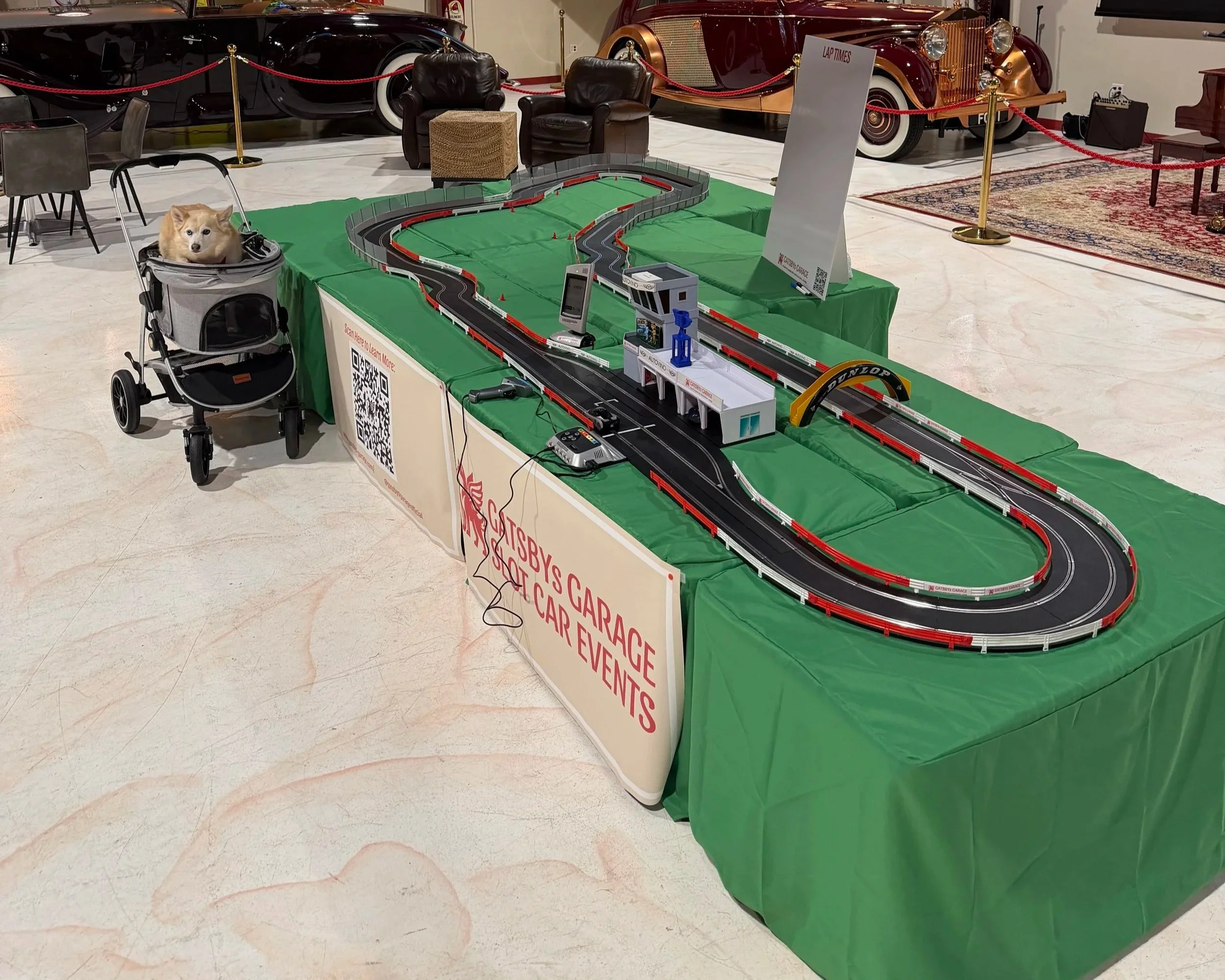 A miniature racing track set up on a table with red and black lanes, a small finish arch, and various racing accessories. There are two black leather armchairs, a small ottoman, a sign for lap times, and classic cars displayed in the background behind a red rope barrier. A dog in a stroller is positioned nearby.