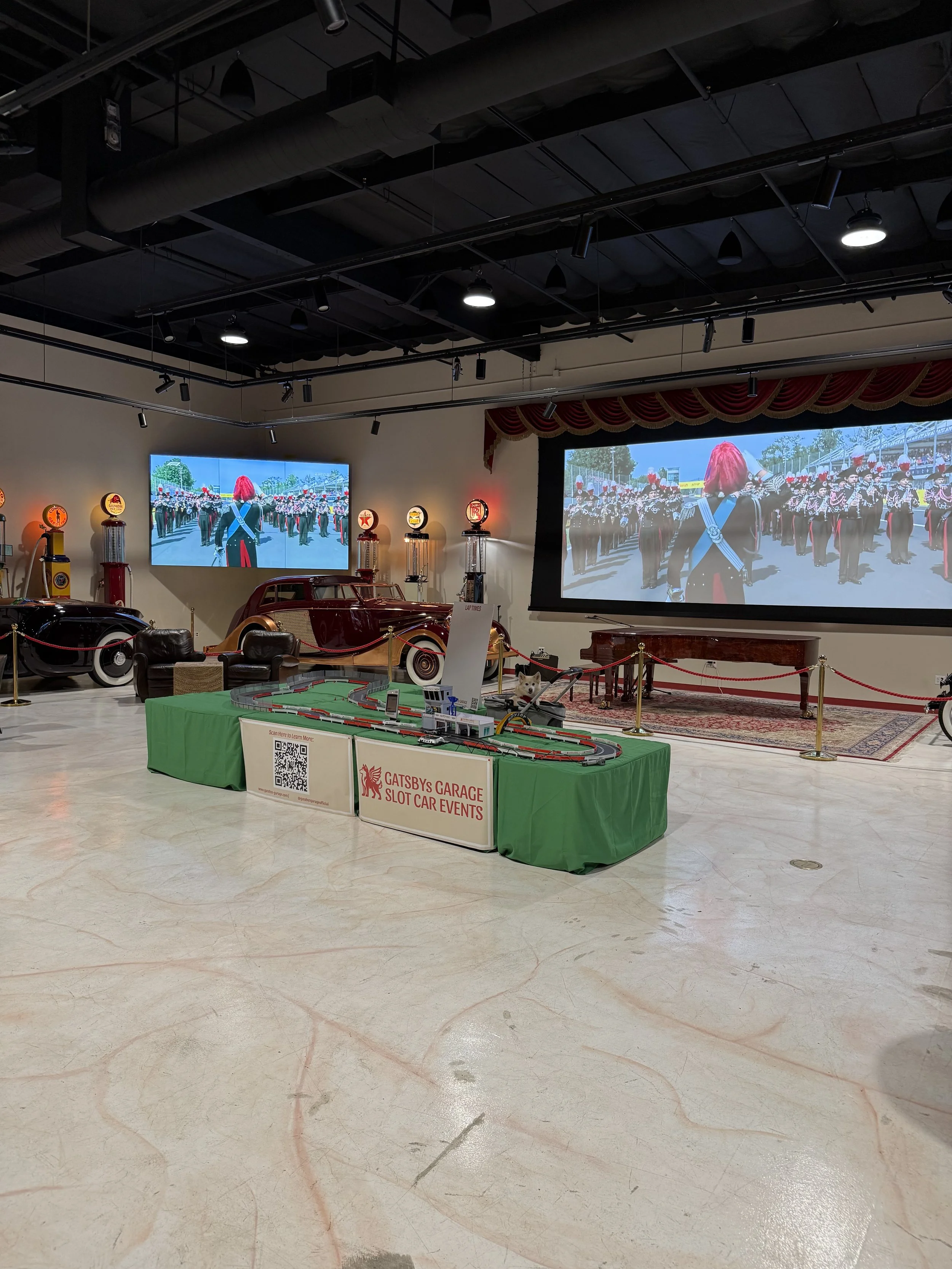 An indoor exhibit at Gatsby's Garage Slot Car Events with two large screens showing a parade of musicians in uniform and red hats. The display includes vintage cars, a model race track, and memorabilia. The floor is marble with visible red and black streaks.