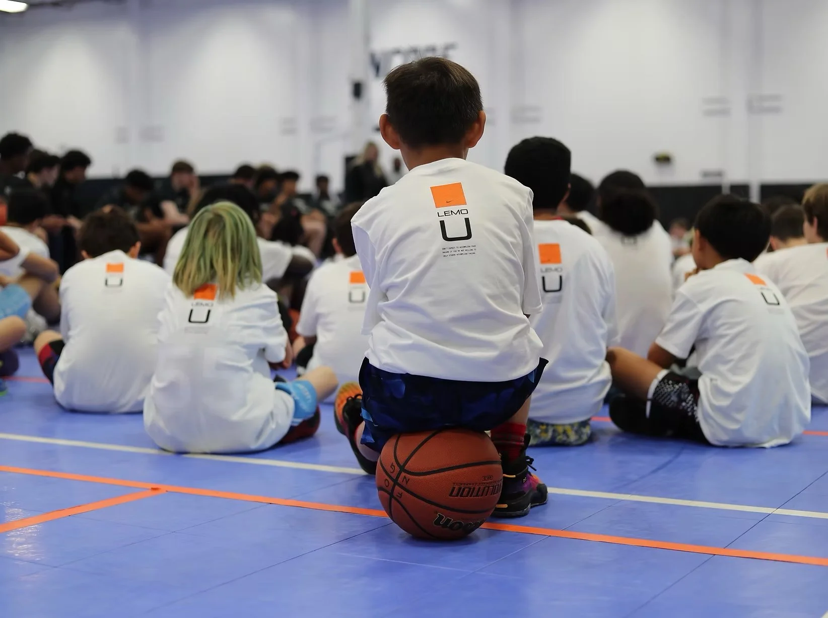 Young children sitting cross-legged on a gym floor during a sports camp or training session, with a young boy in the foreground kneeling with a basketball underneath him, in a gymnasium.