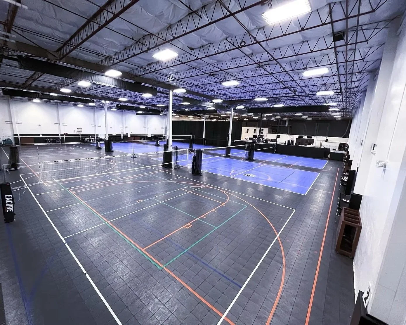 Indoor sports facility with multiple badminton courts, each marked with colorful lines, and netting separating them. The ceiling is high with industrial lighting and structural beams. There are some equipment and a few people working at computers in the background.