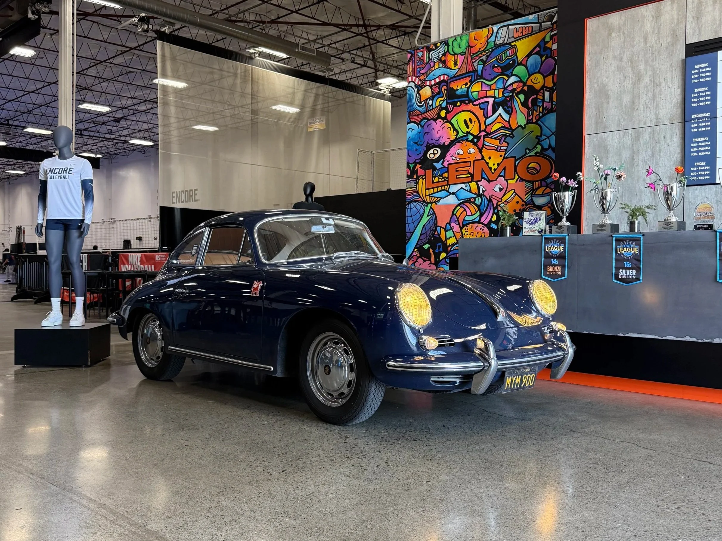 A vintage black Porsche car with a yellow license plate labeled 'MYM 900' parked indoors, with a mannequin wearing sportswear and a graffiti-style colorful mural labeled 'LEMO' in the background, and trophies on a table to the right.