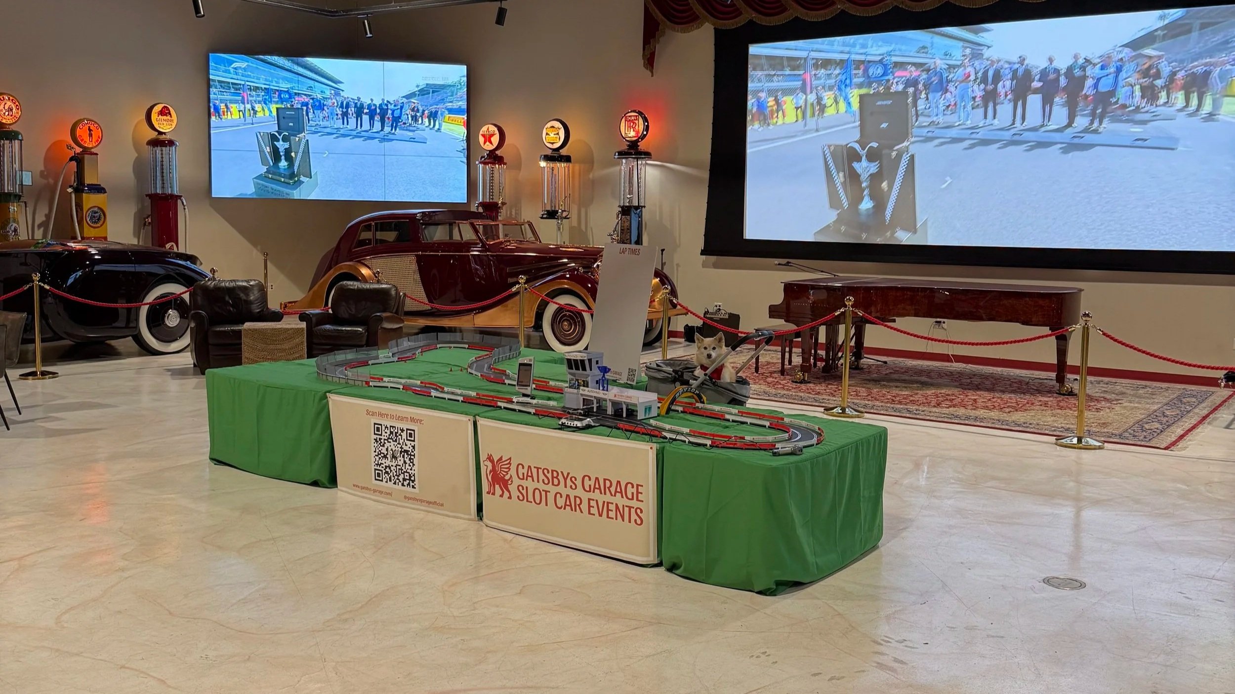 This image shows a vintage car exhibit with a model train setup on a green table in the foreground, and two large screens displaying a racing scene. Classic cars are on display in the background, and there are vintage gas pumps and decorative signs on the wall. A sign on the table indicates the event is hosted by Gatsby's Garage Slot Car Events.