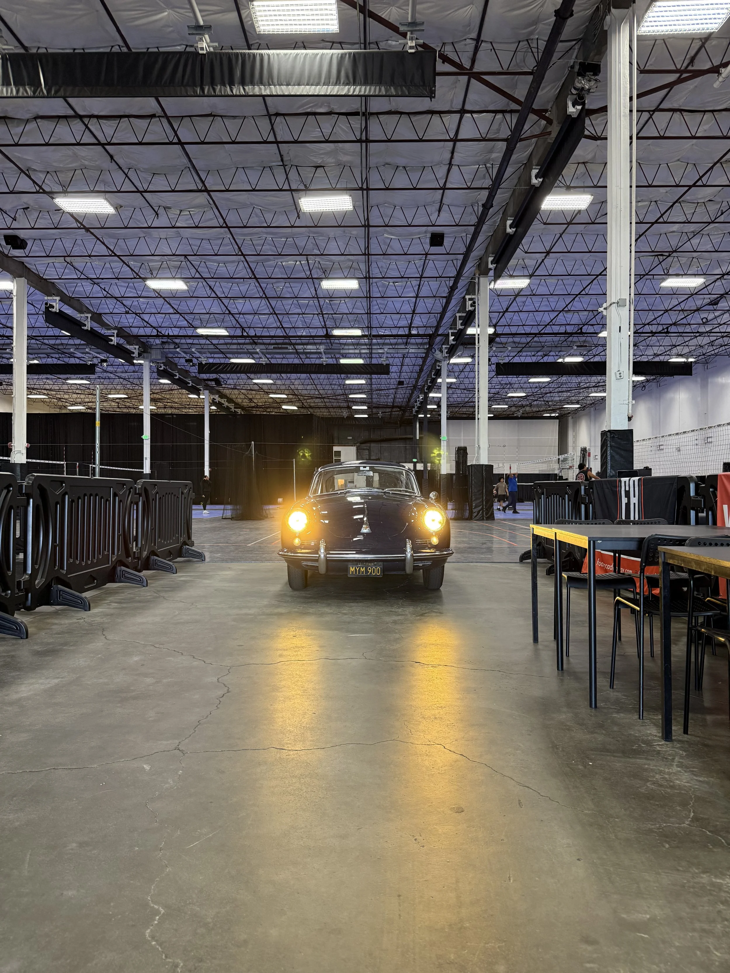 An indoor volleyball court with tables, chairs, and a vintage black car with glowing headlights in the center.