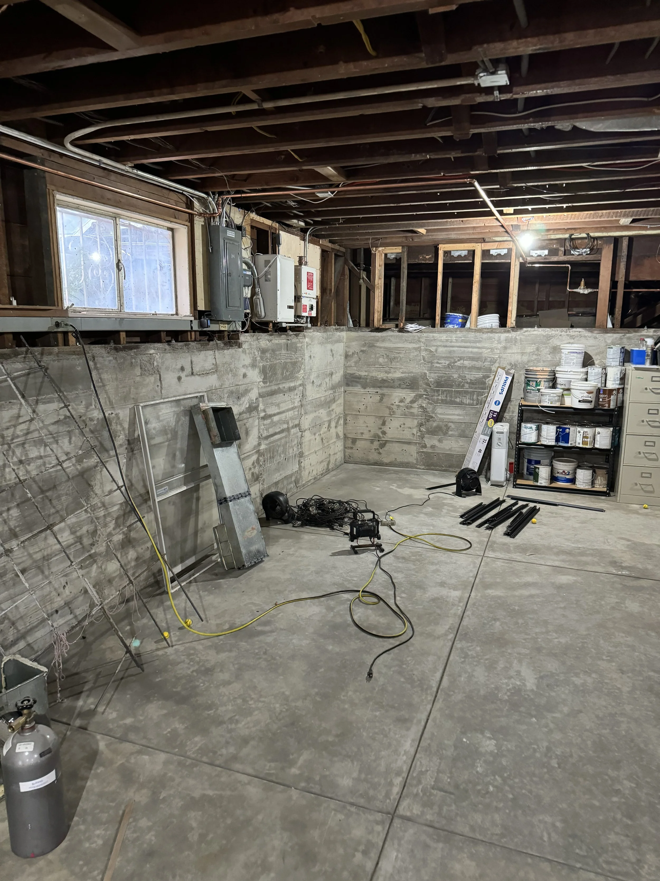 Empty basement with exposed wooden beams, concrete walls and floor, scattered tools and paint cans, shelving, and wiring on the floor and ceiling.
