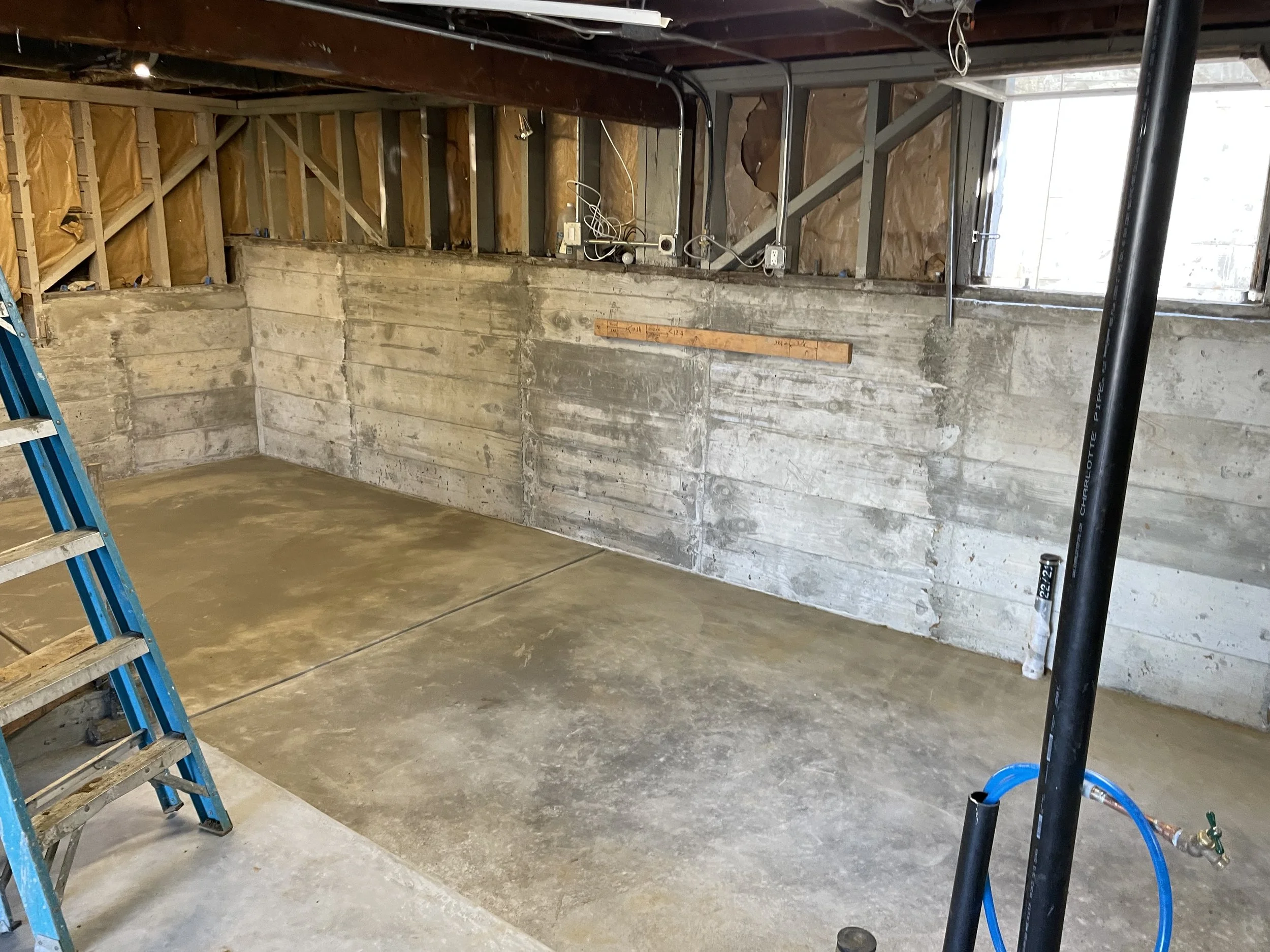 Unfinished basement with exposed insulation and concrete walls, a blue ladder, and exposed pipes.