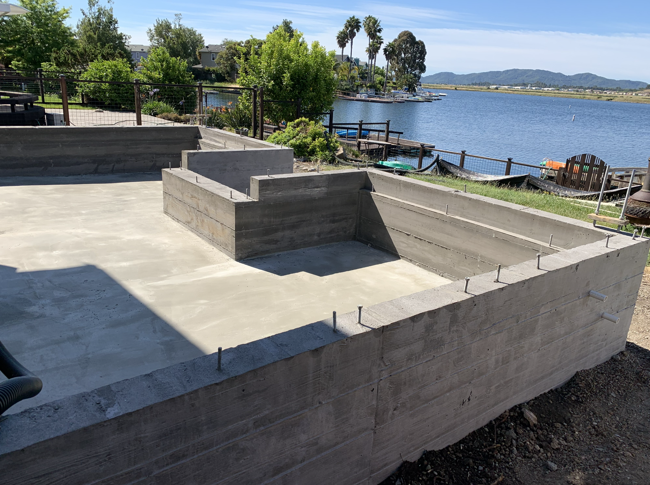 Concrete foundation of a structure near a lake, with a flat surface and some vertical protrusions. Trees and a fenced area by the water in the background.
