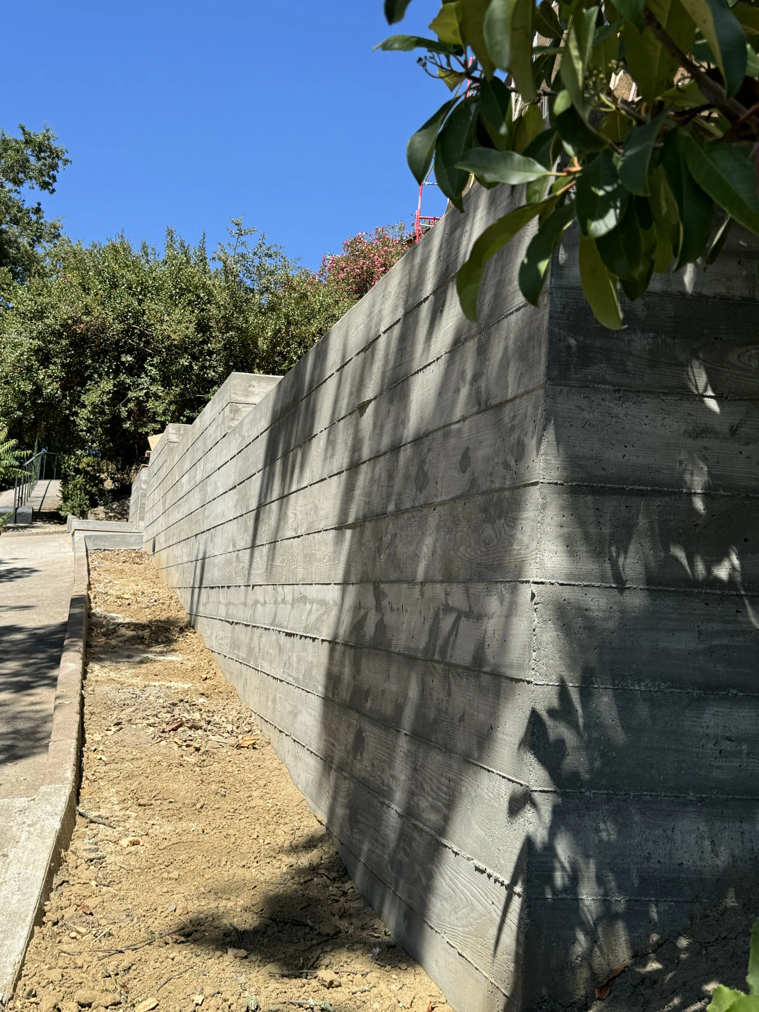 Concrete retaining wall with shadows from trees, surrounded by soil and greenery, under a clear blue sky.