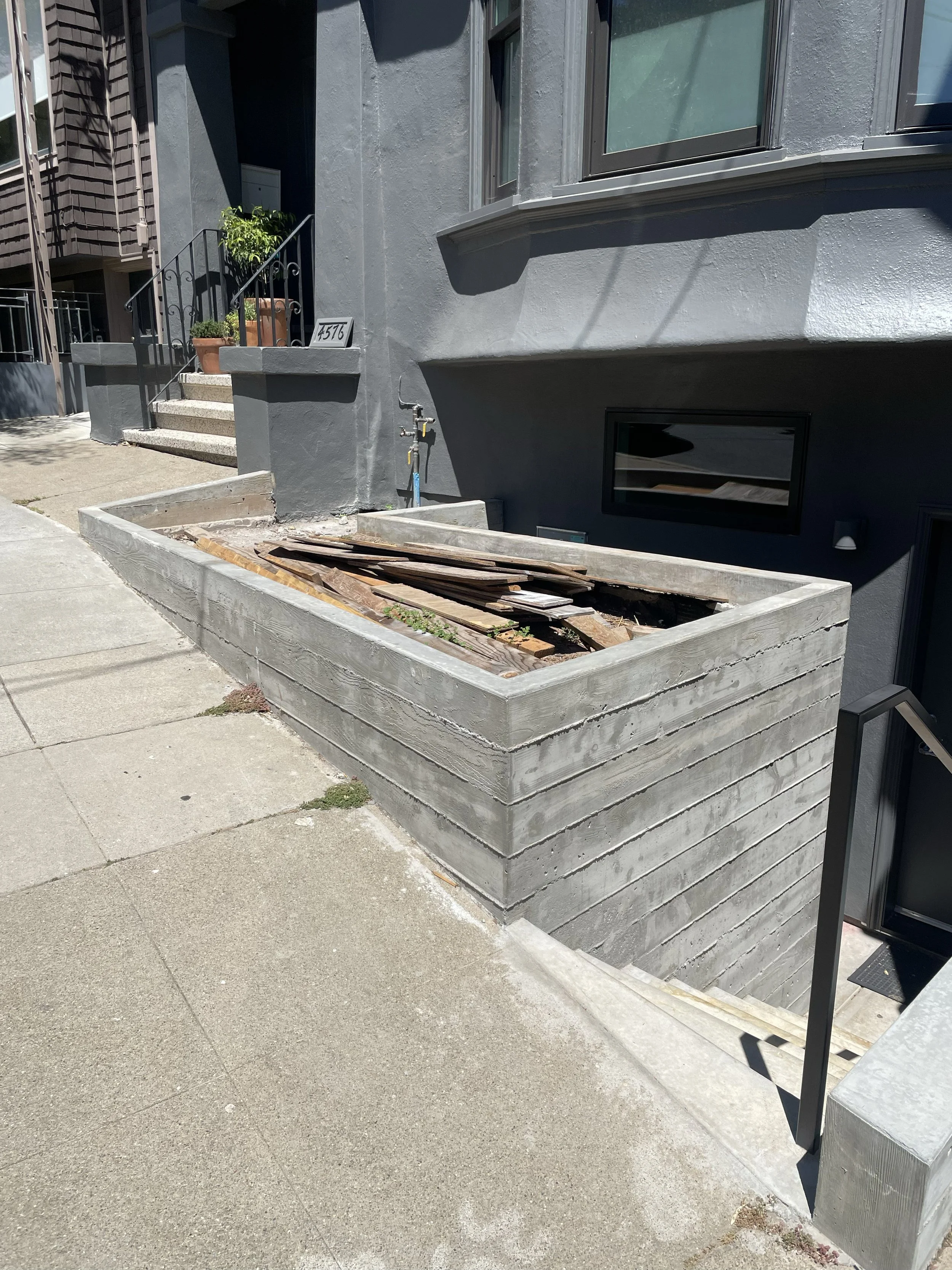 Concrete planter on walkway with wood inside near apartment entrance.