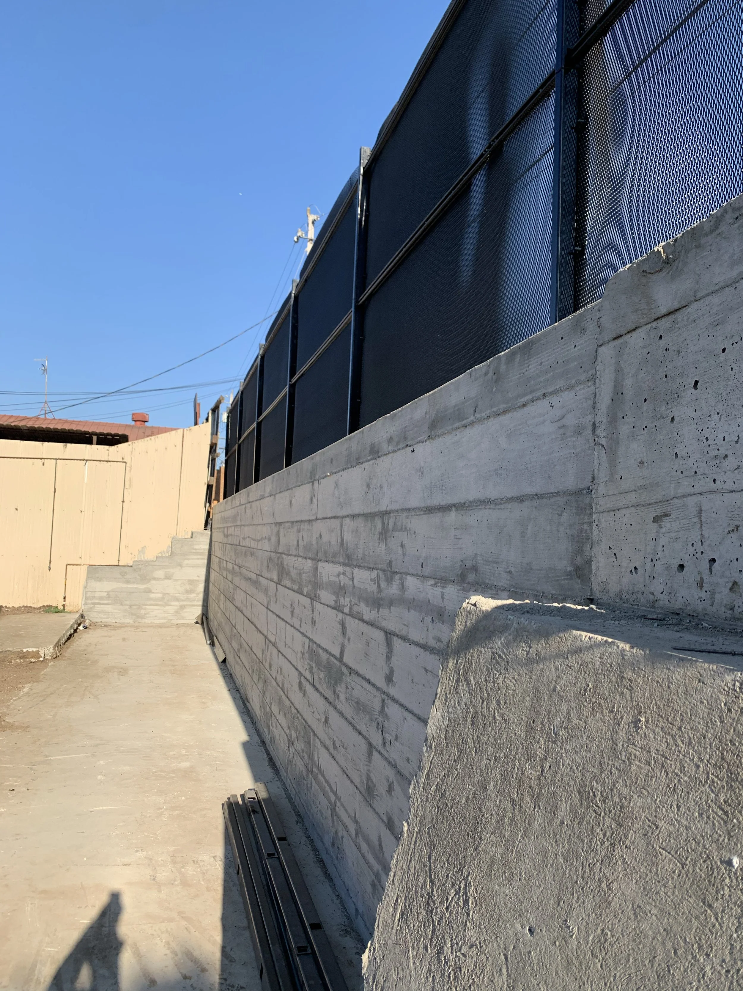 Concrete wall with metal fence on top, adjacent to a sloped pavement and wooden structure, under clear blue sky.