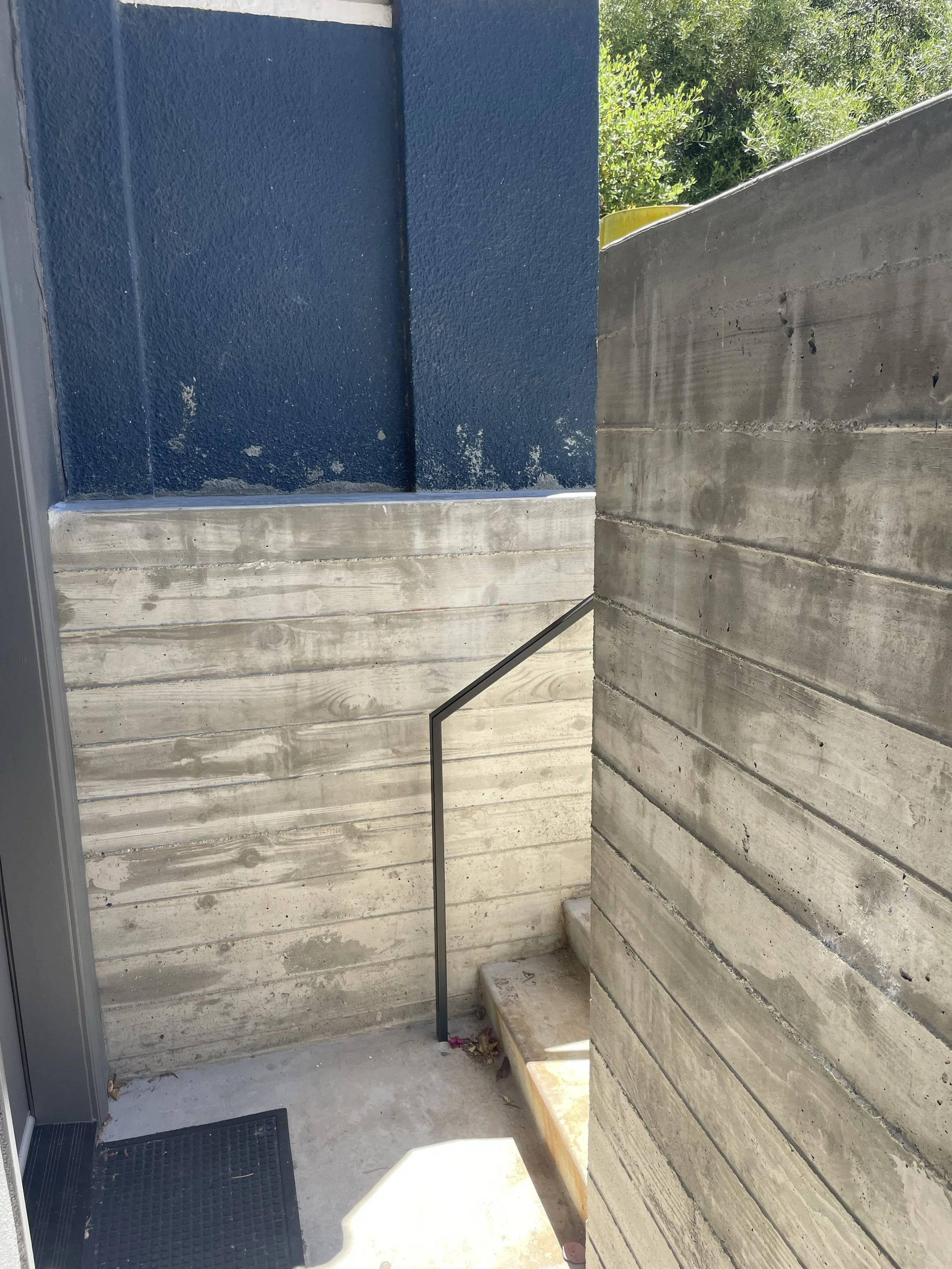 Concrete steps and walls with a black metal railing next to a blue building exterior, partially in sunlight.