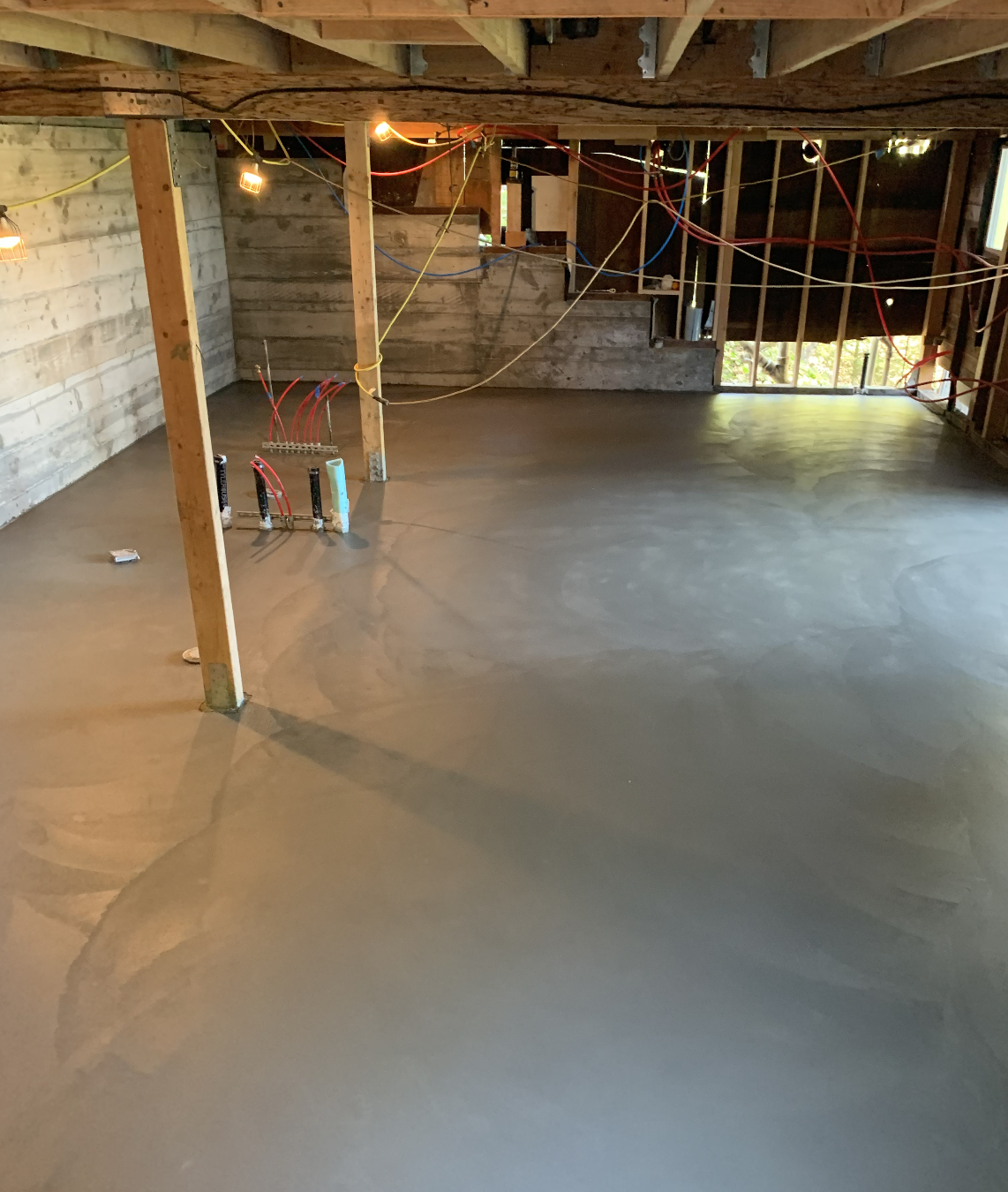 Concrete basement floor with wooden support beams, visible wiring, and plumbing pipes.