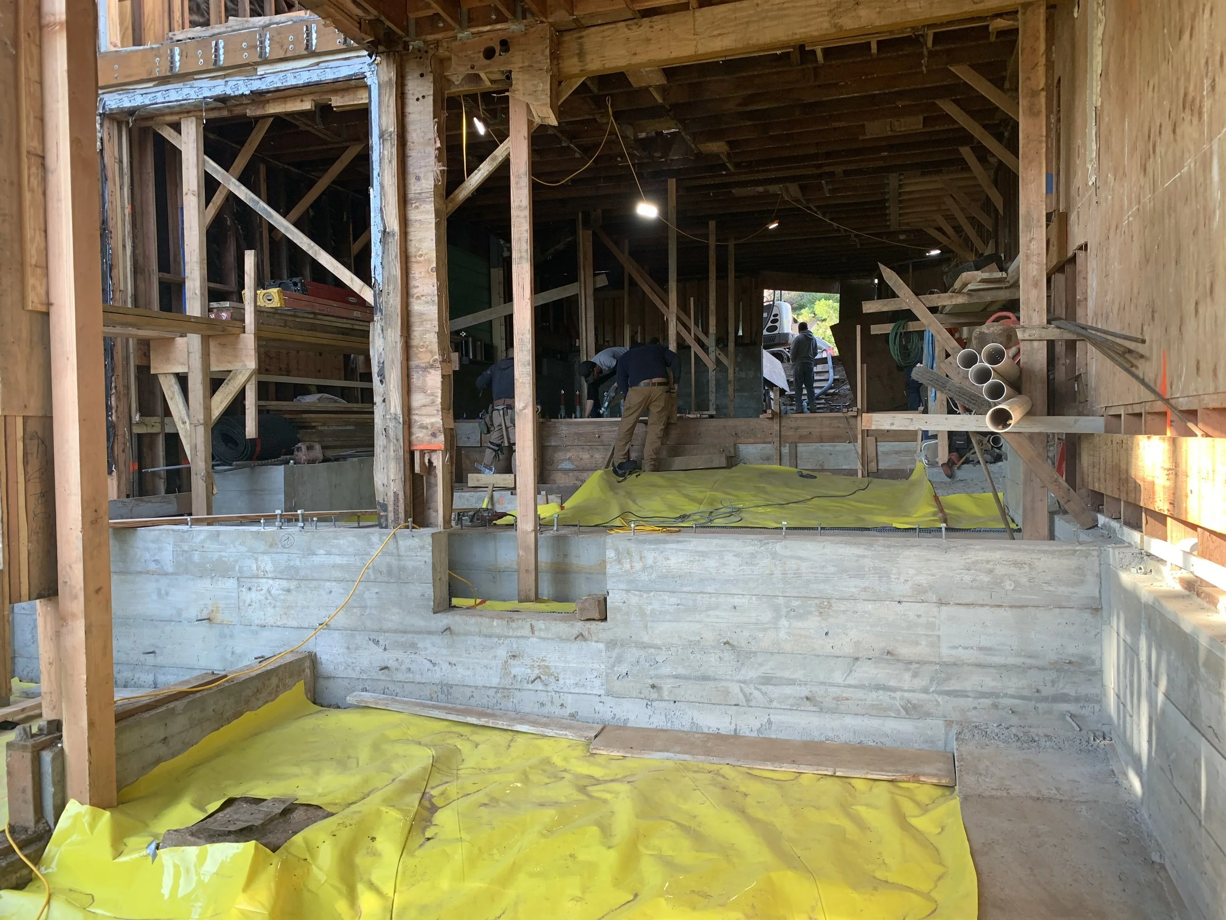 Construction site interior with wooden framework, yellow tarp on concrete floor, and workers in the background.