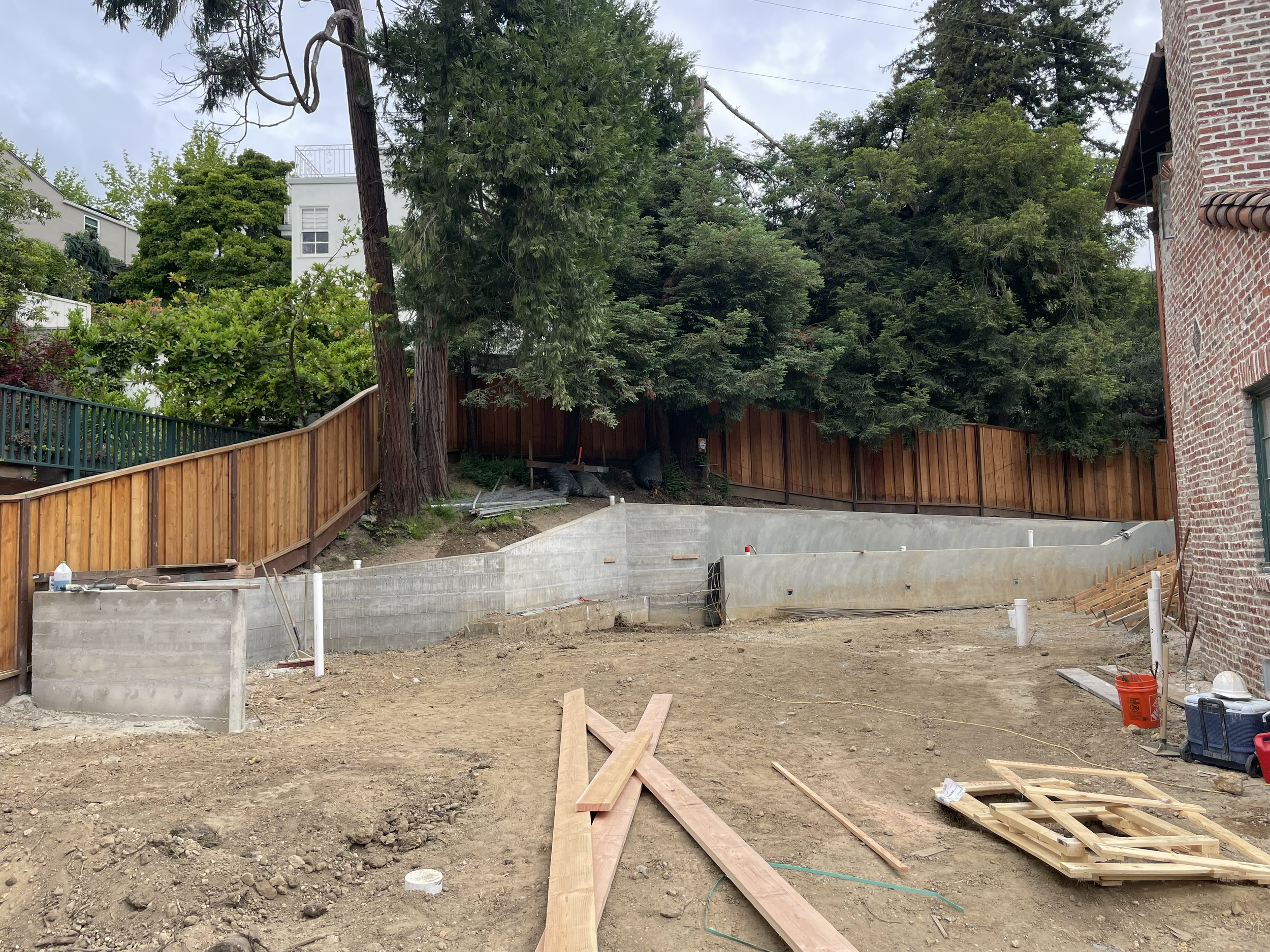 Construction site with wooden planks, dirt ground, concrete walls, and wooden fence with trees around.