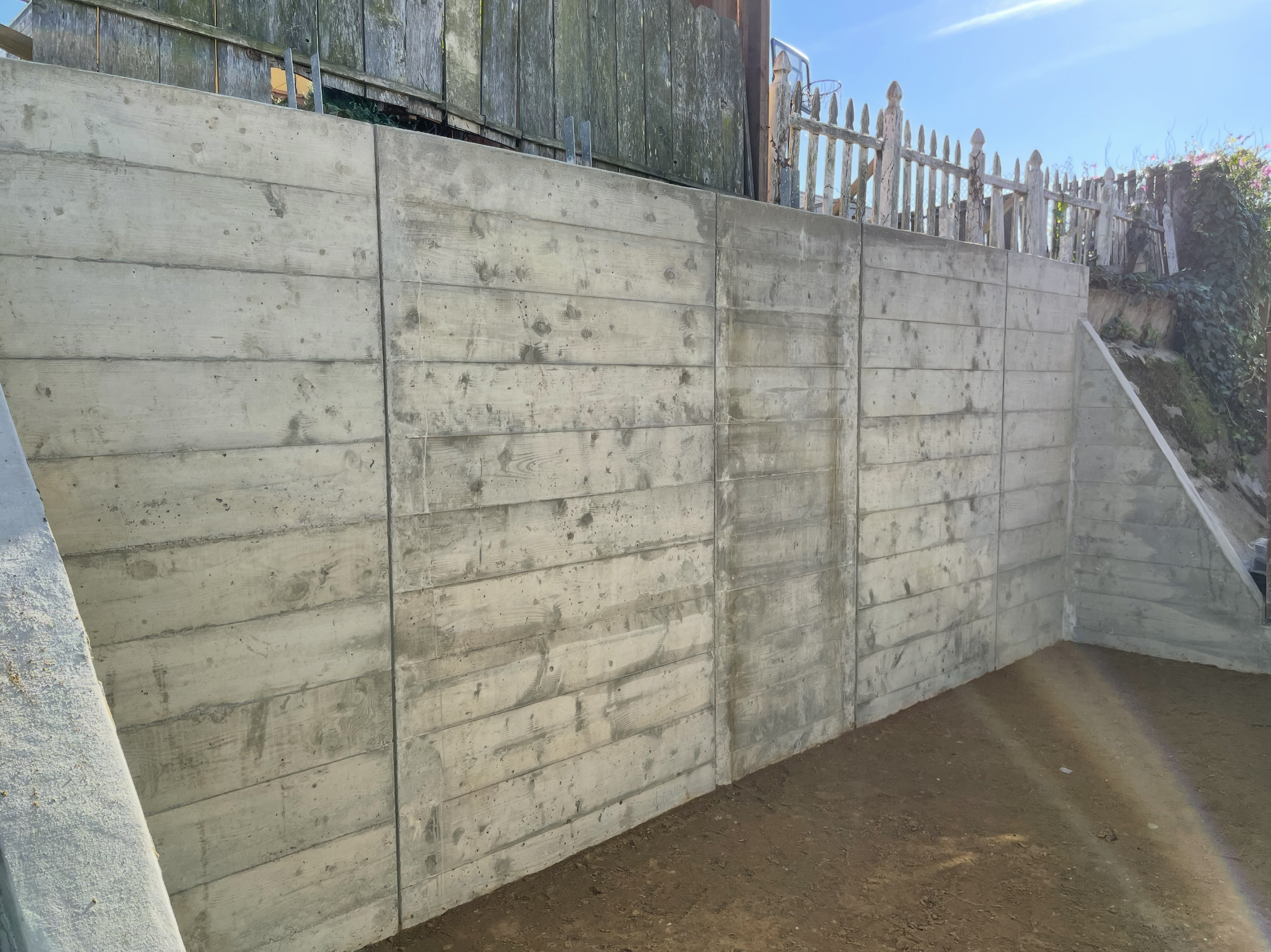 Concrete retaining wall with wooden fence on top, blue sky background.