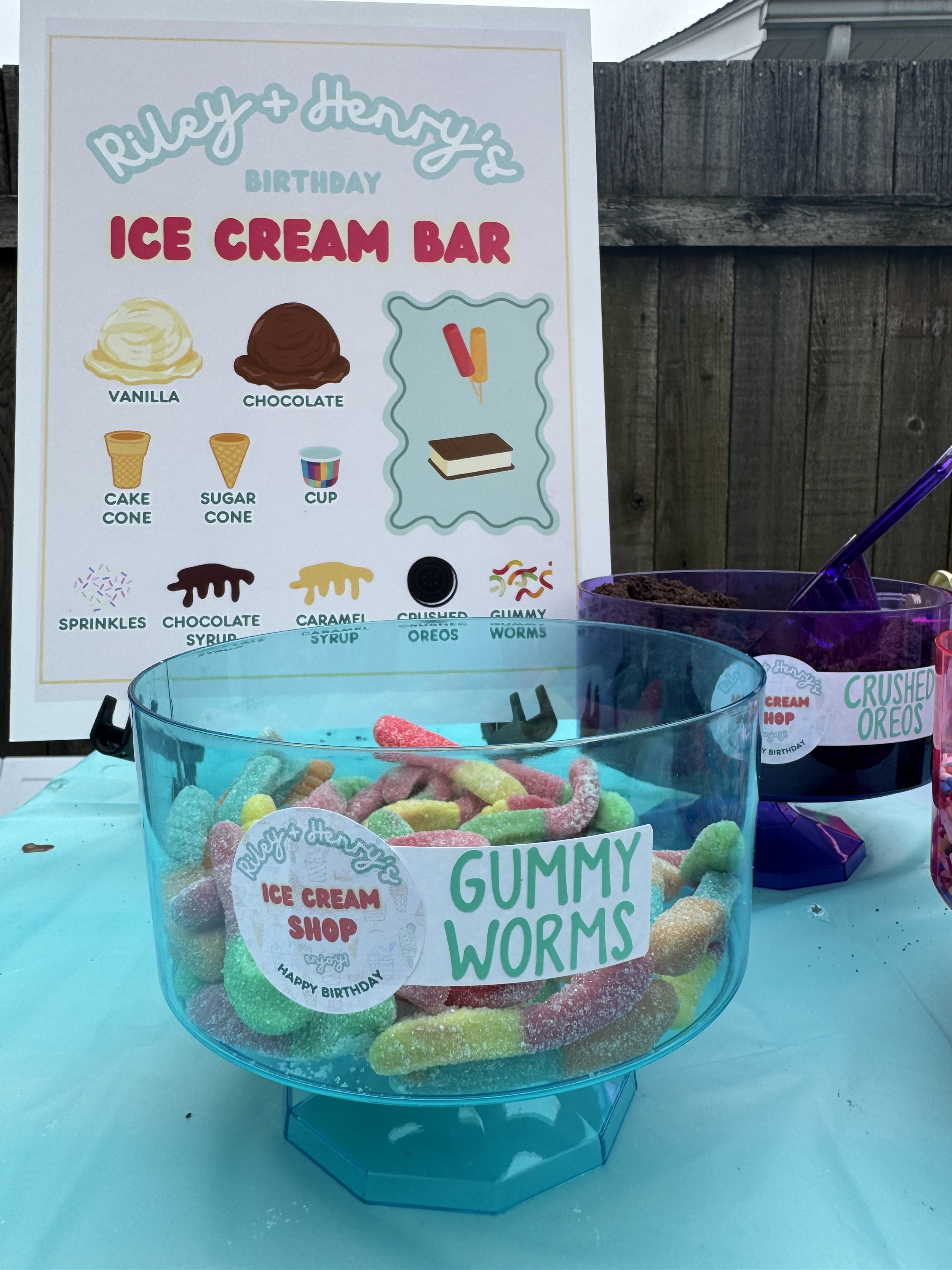 A container of colorful gummy worms at a birthday party setup with a sign and other candy containers in the background.