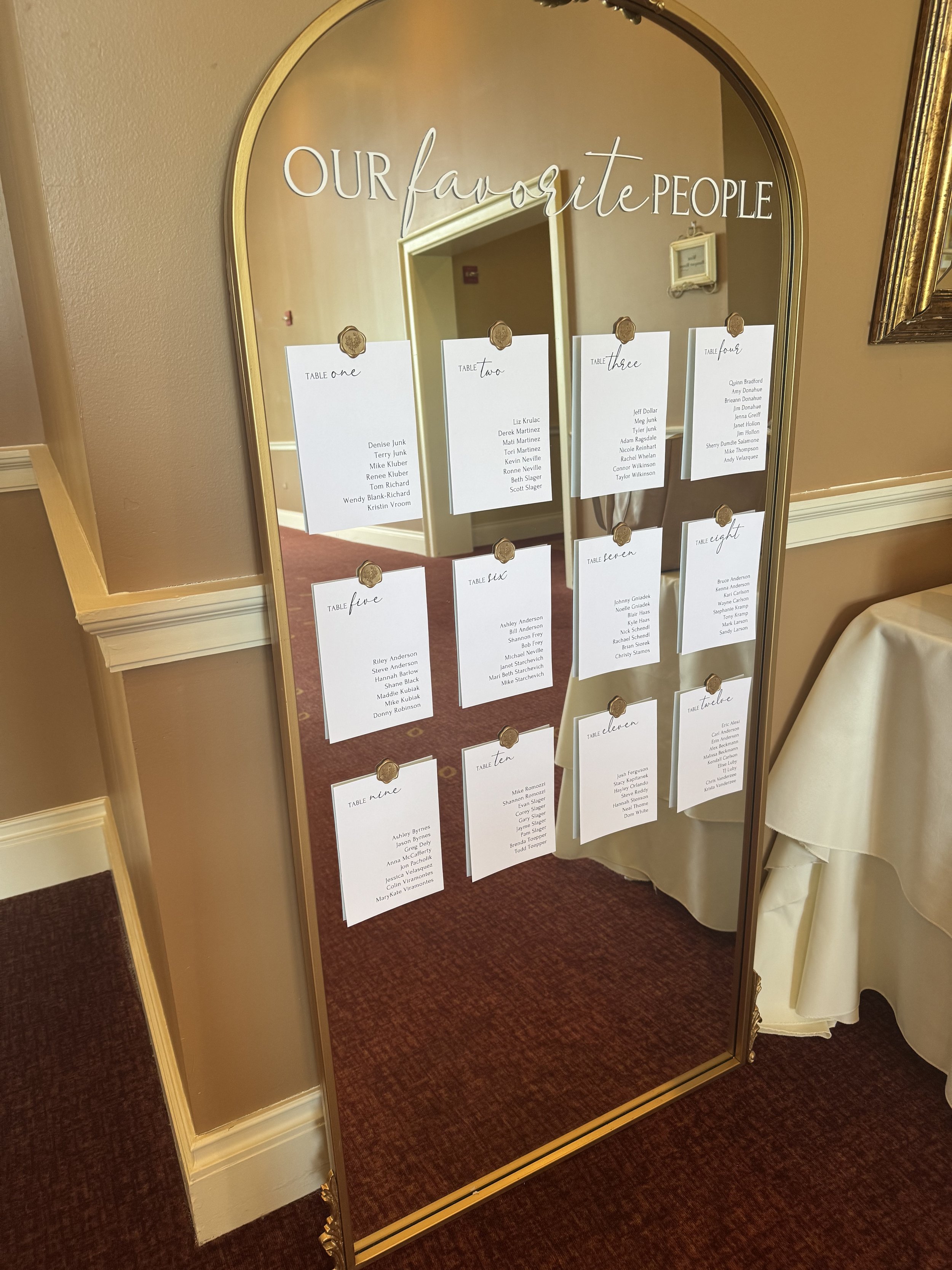 A large mirror with gold trim displays wedding seating arrangements, titled 'Our Favorite People,' with printed lists of names organized by table number, attached with gold pins.