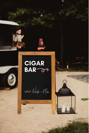 Chalkboard sign reading 'Cigar Bar' with additional text and arrow, outdoor setting with trees, lantern, candles, and a golf cart in background.