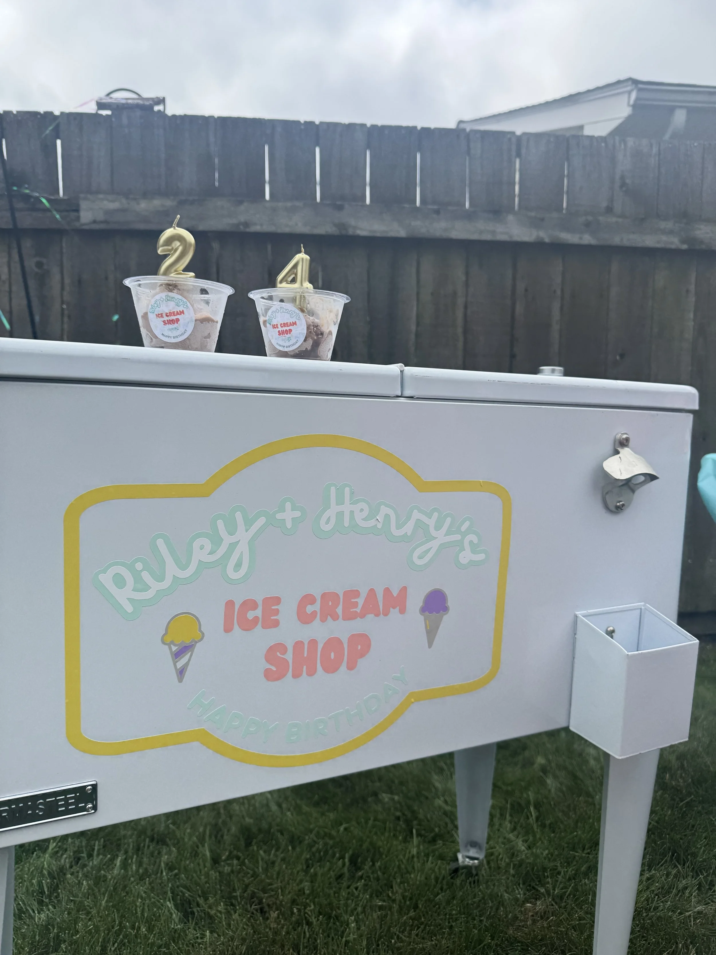 Ice cream shop sign with colorful lettering and small ice cream cone graphics, two cups of ice cream with candles shaped as the numbers 2 and 4 on top placed on an outdoor white freezer, fenced backyard with cloudy sky in the background.