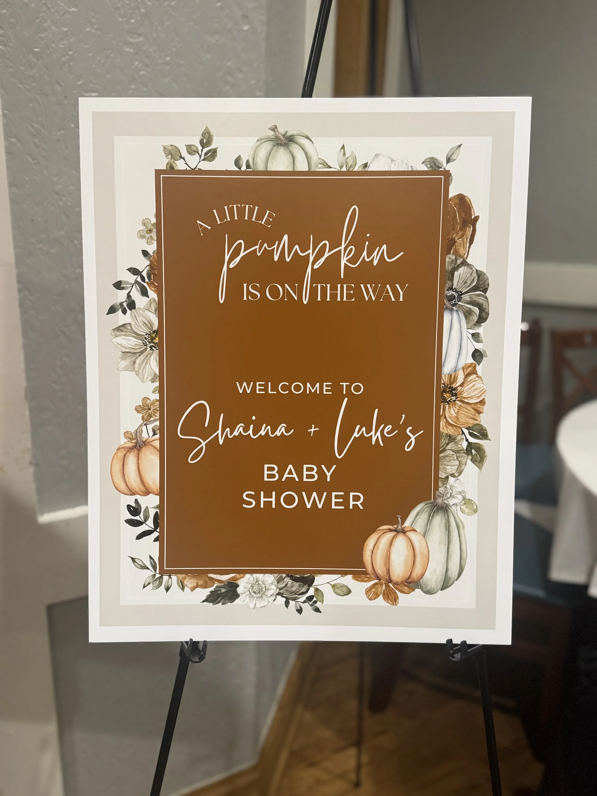 Welcome sign for Shaina and Luke's baby shower with a fall pumpkin and floral border.