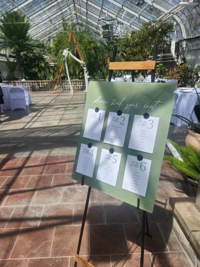 A seating arrangement sign for a wedding or event, indicating table numbers 1 through 6 with guest names, placed inside a greenhouse or conservatory with glass ceiling and lush greenery.