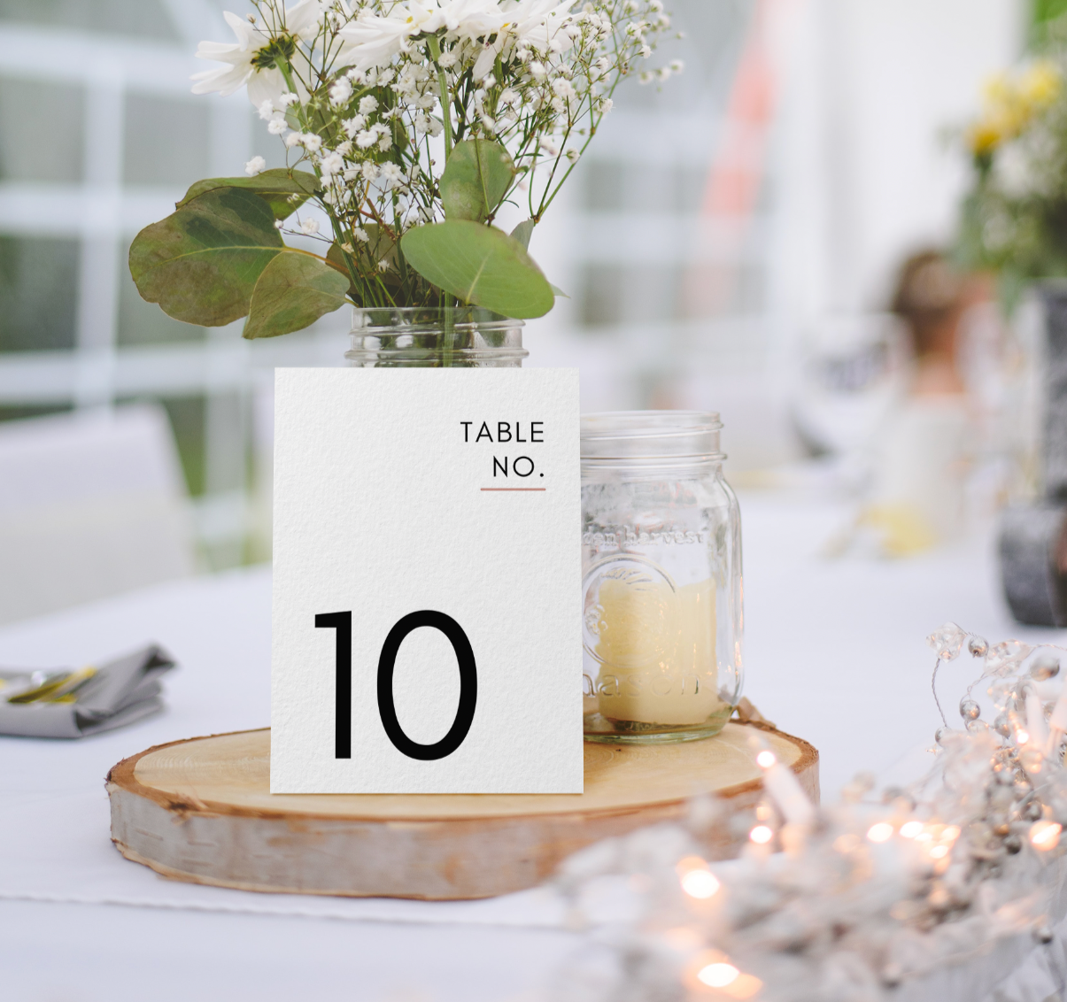 Table number card with the number 10, placed on a wooden slab; a mason jar with white flowers and green leaves, and a glass jar with a candle visible on a white tablecloth at a decorated event.