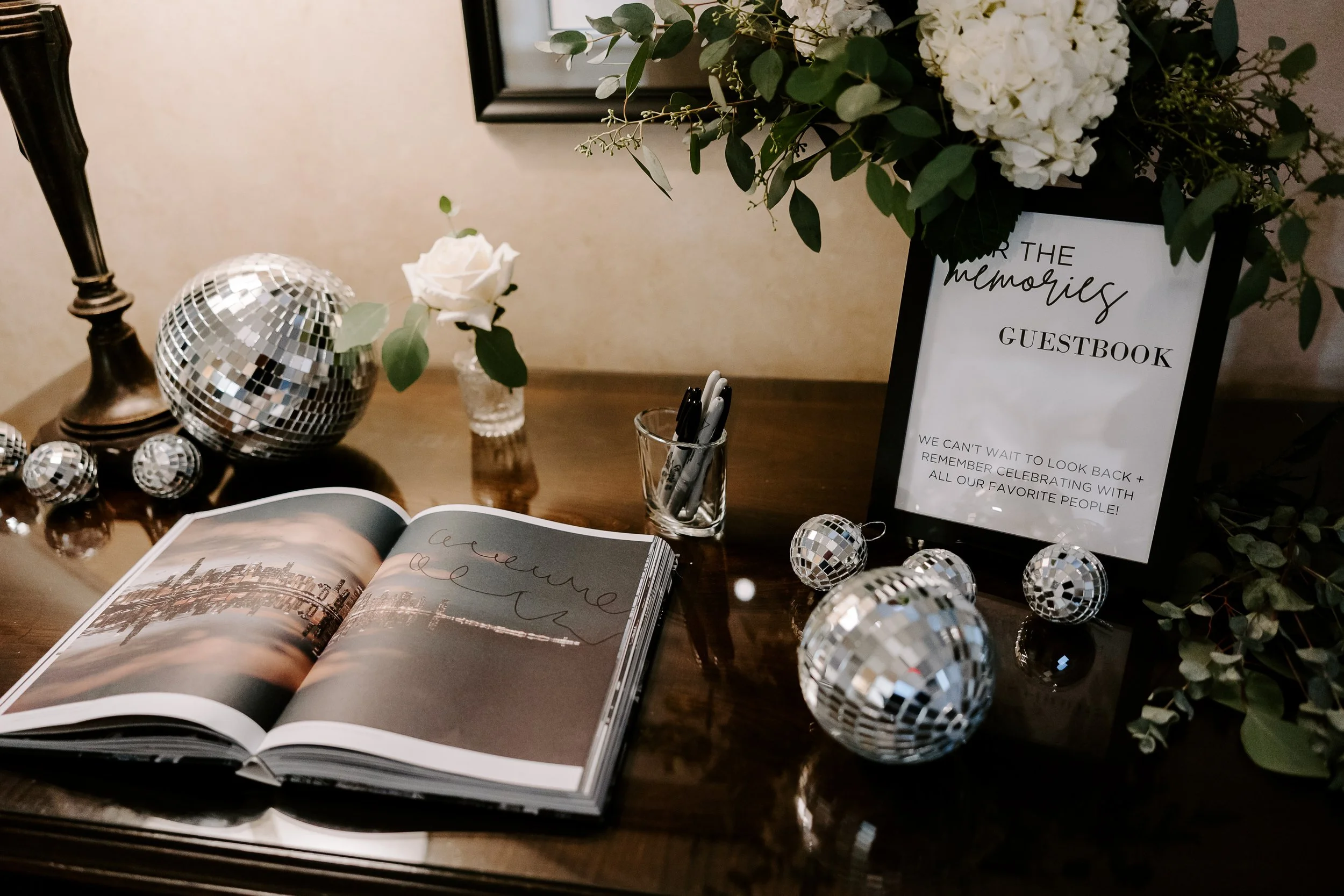 A guestbook on a wooden table with a photo of a cityscape, surrounded by disco ball ornaments, a vase with a white rose, a sign that says 'For the Memories Guestbook,' a flower arrangement with white flowers, and a small container with black and white pens.