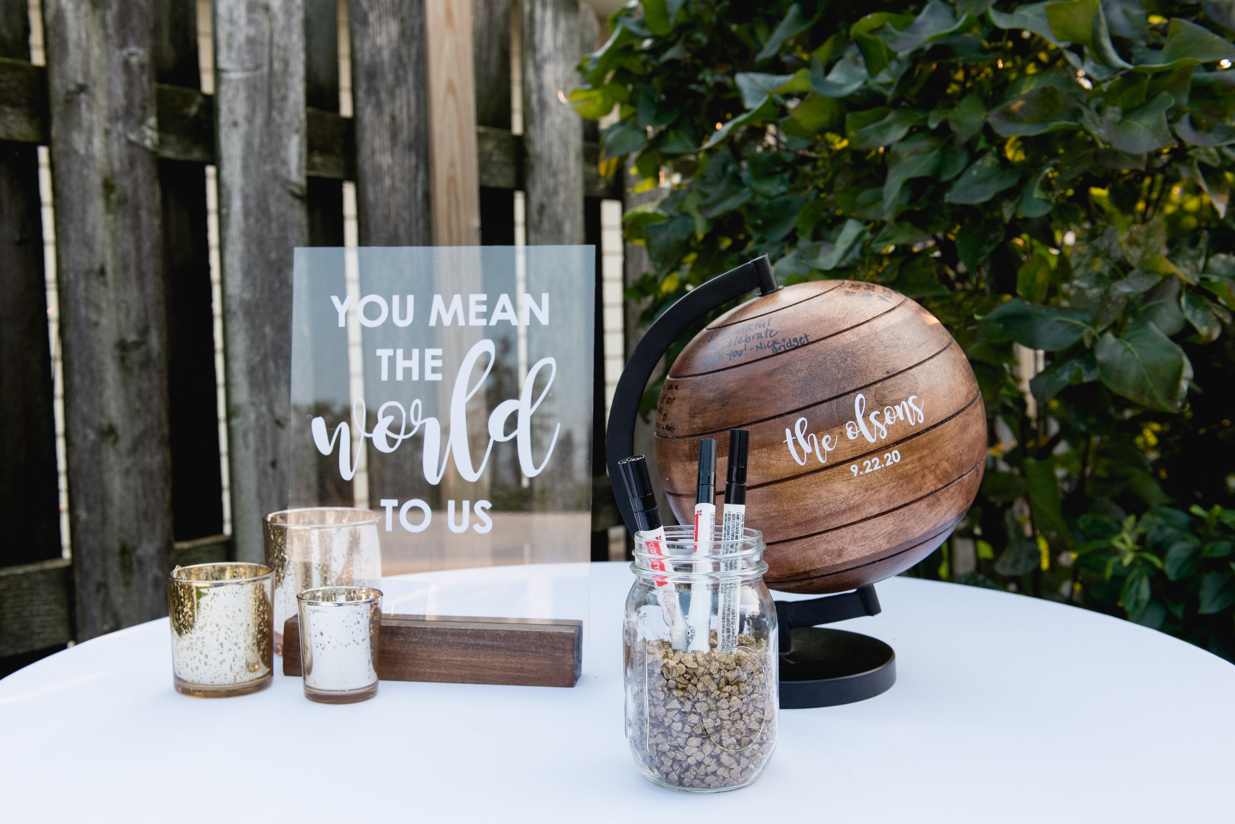 Decorative table with a wooden globe with writing and date, a glass jar filled with markers, three small candles with gold speckles, and a sign that says "You mean the world to us" in an outdoor setting with a wooden fence and leafy bushes