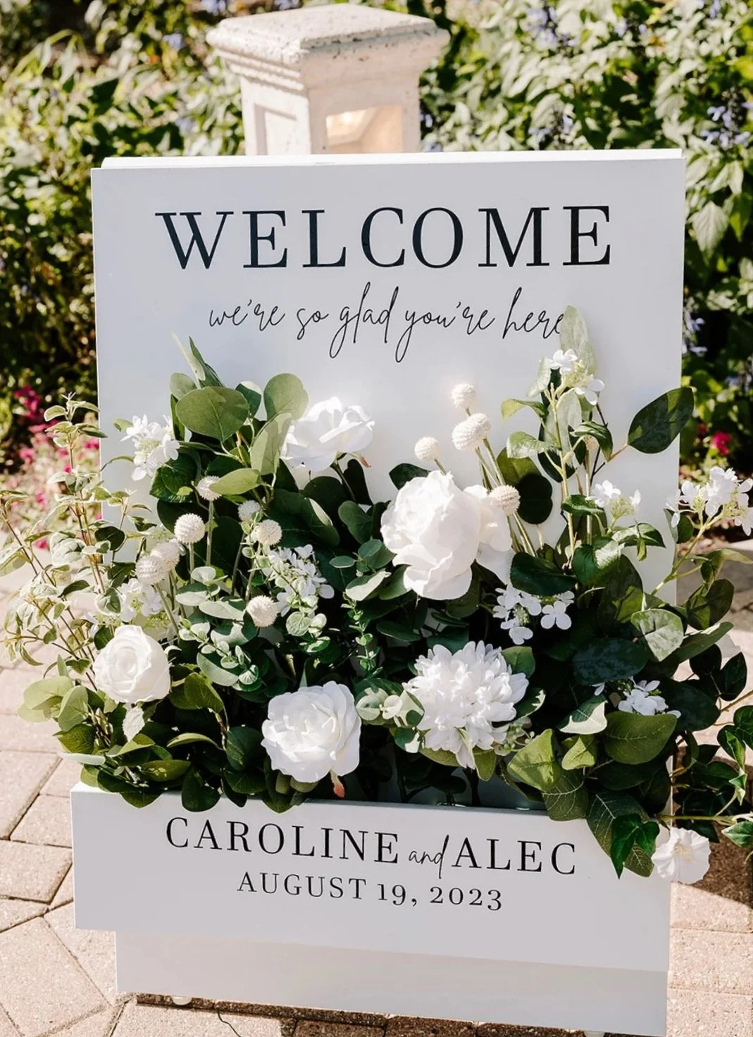 A wedding welcome sign with white flowers and green leaves, displaying a greeting and wedding details.