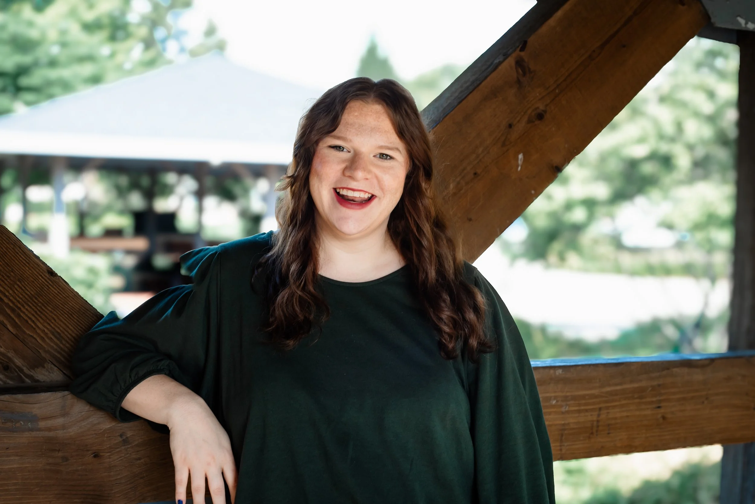 Woman with brown hair in a green top, smiling on a bridge for a headshot.