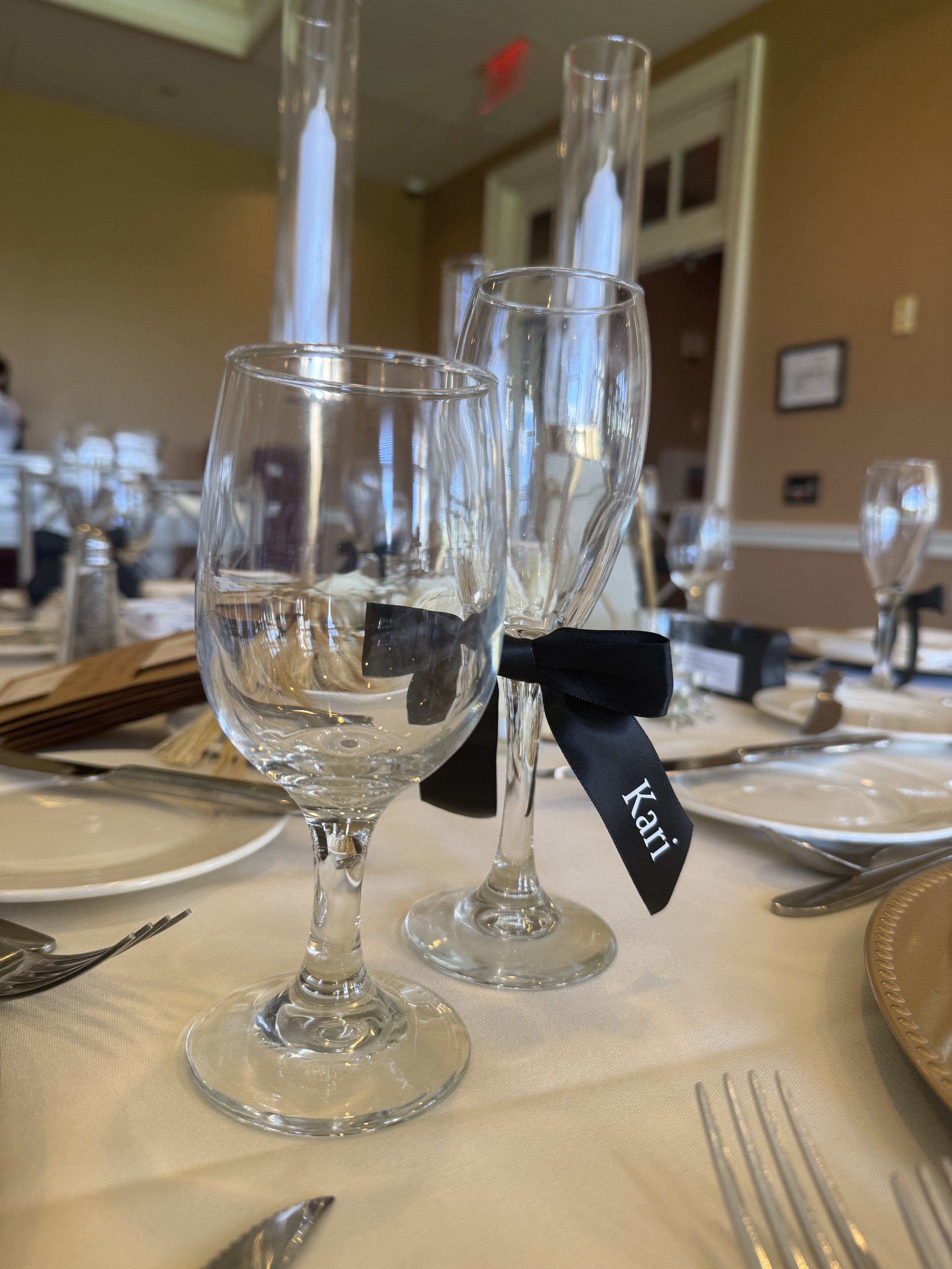 Table setting with a water glass, champagne flute with a black ribbon labeled 'Kai', and additional glassware, plates, and utensils at a formal event.