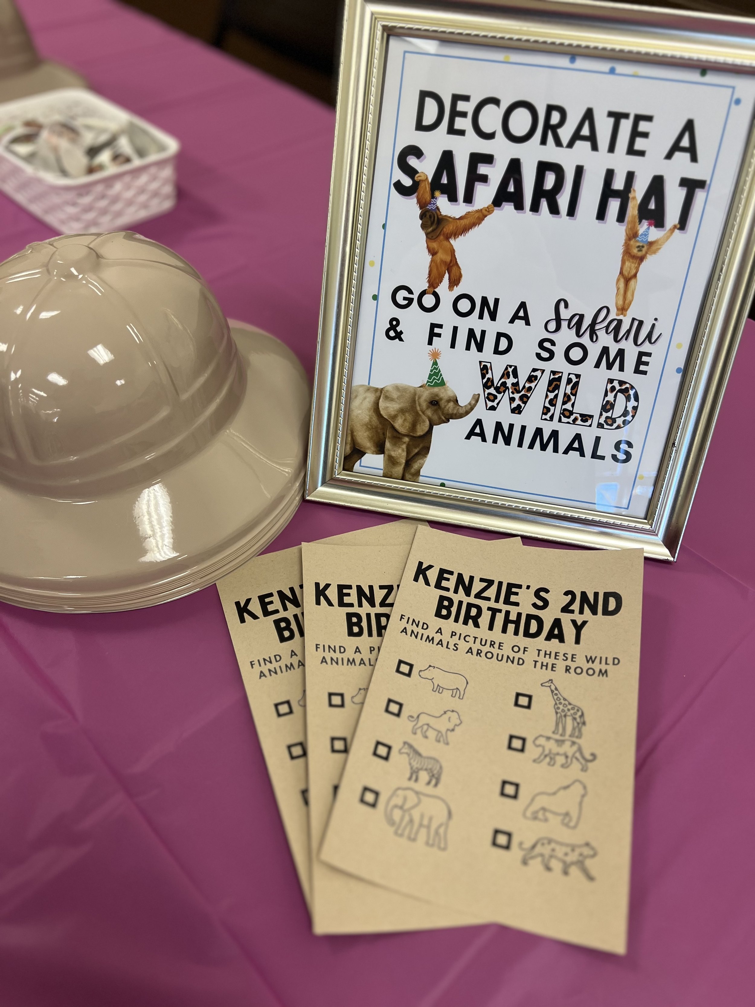 Party table with a framed sign promoting a safari hat decorating activity, showing cartoon animals wearing party hats, and paper activity sheets titled "Kenzie’s 2nd Birthday" with pictures of wild animals for finding around the room.