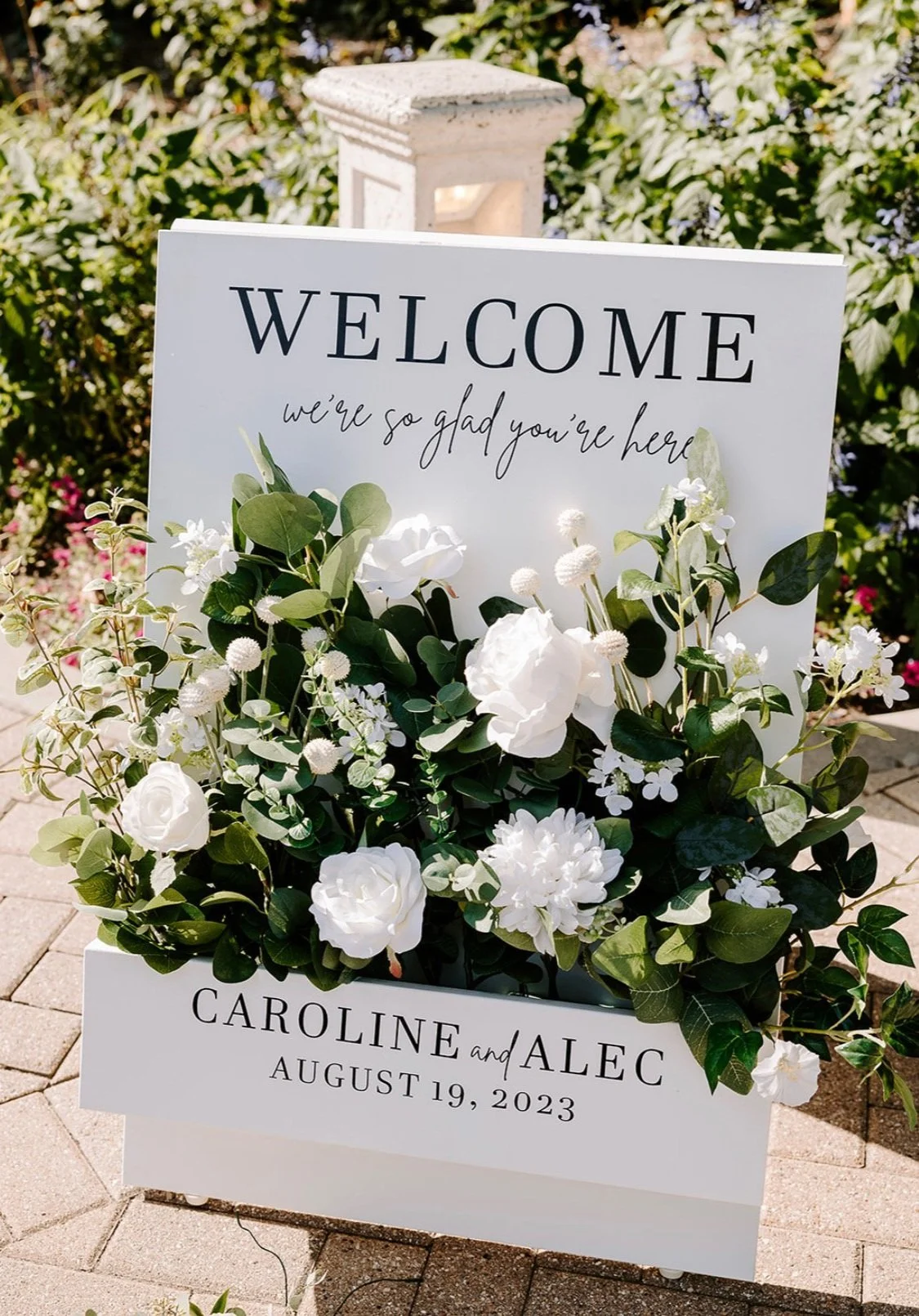 Wedding welcome sign with white flowers and green foliage, displaying the message 'Welcome, we're so glad you're here.' It features the names Caroline and Alec and the date August 19, 2023.