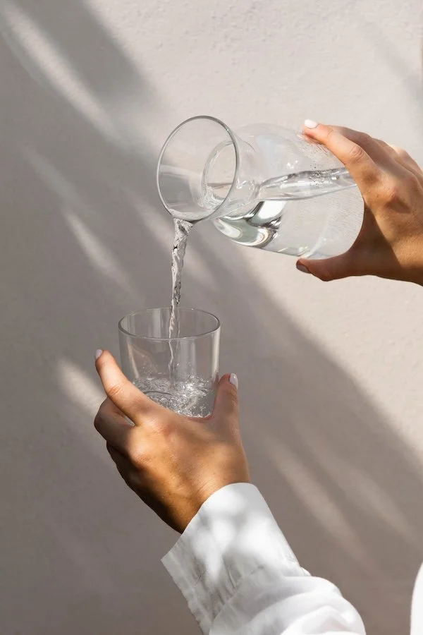 Person pouring water from a glass pitcher into a glass on a light background.