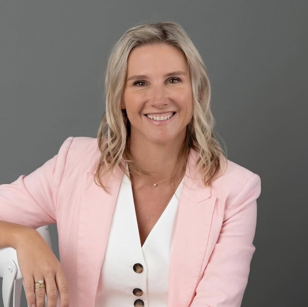 Smiling woman with blonde hair wearing a pink blazer and white top, seated against a gray background.