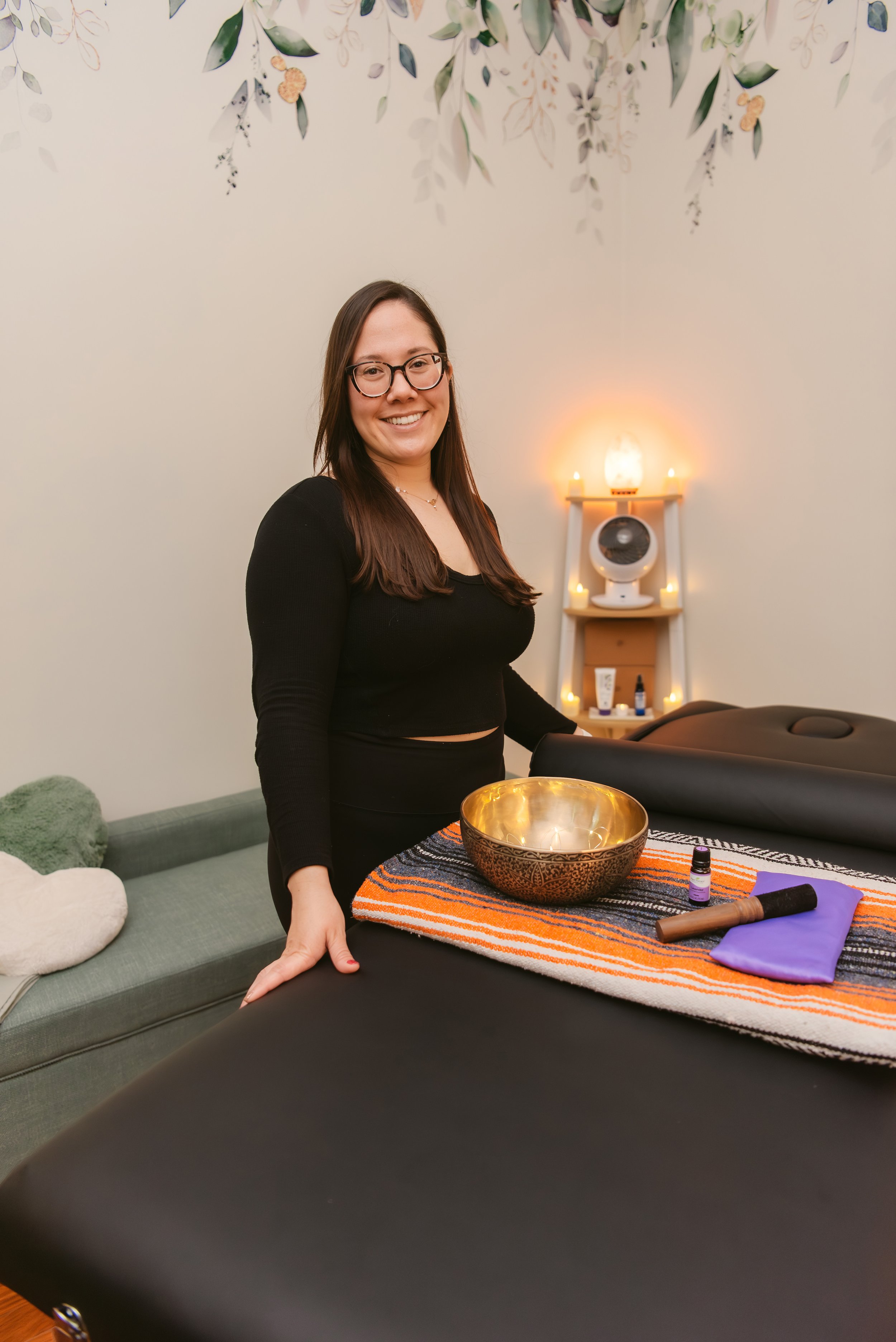A woman with long dark hair and glasses smiling while standing next to a massage table with a singing bowl, a small purple bottle, and other items on a colorful striped cloth. Behind her is a wooden shelf with candles and a salt lamp, and a wall decorated with leaf paintings.
