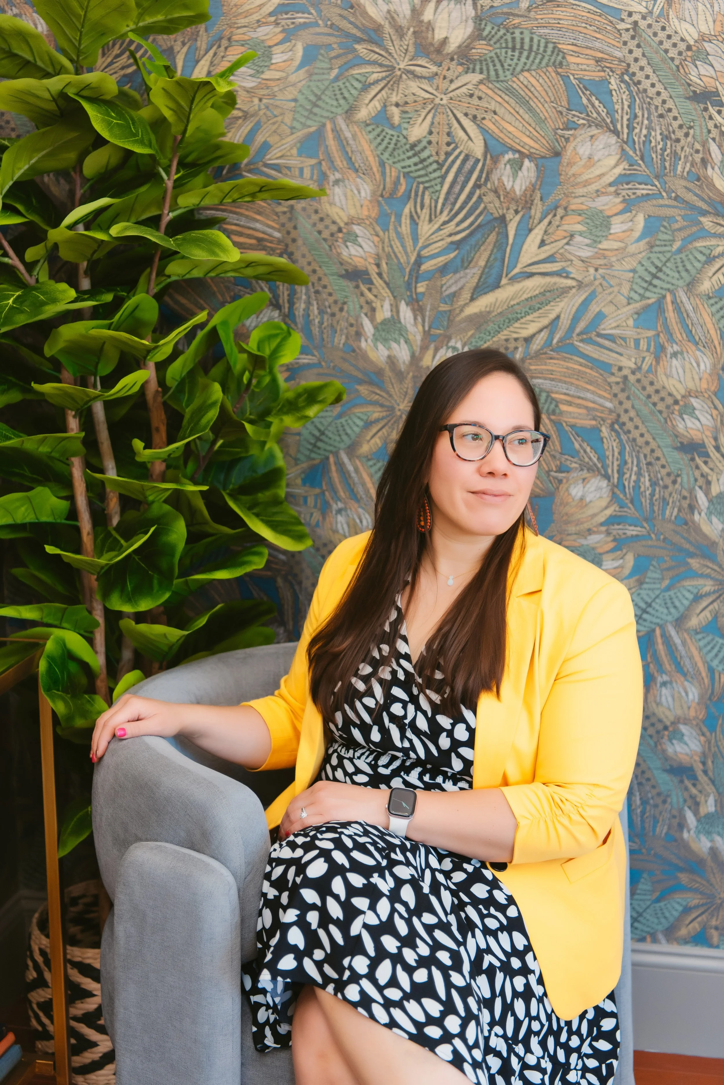 A woman with long dark hair, glasses, and wearing a yellow blazer and black-and-white patterned dress, sitting on a gray armchair. She is inside a room with a large green plant on her left, and a blue patterned wallpaper with tropical leaves behind her.