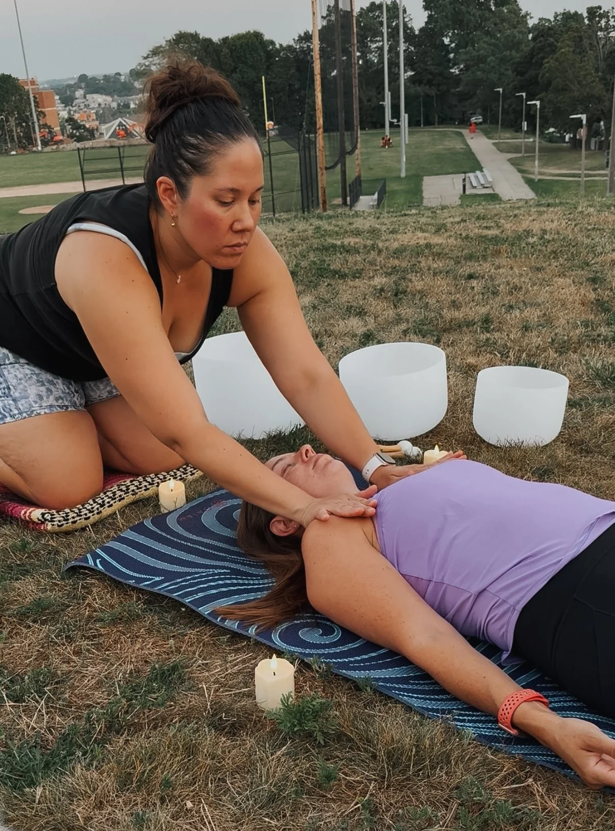 A woman receives a sound healing or relaxation therapy session outdoors on grass, lying on a yoga mat with candles and singing bowls nearby, as another woman performs the therapy.