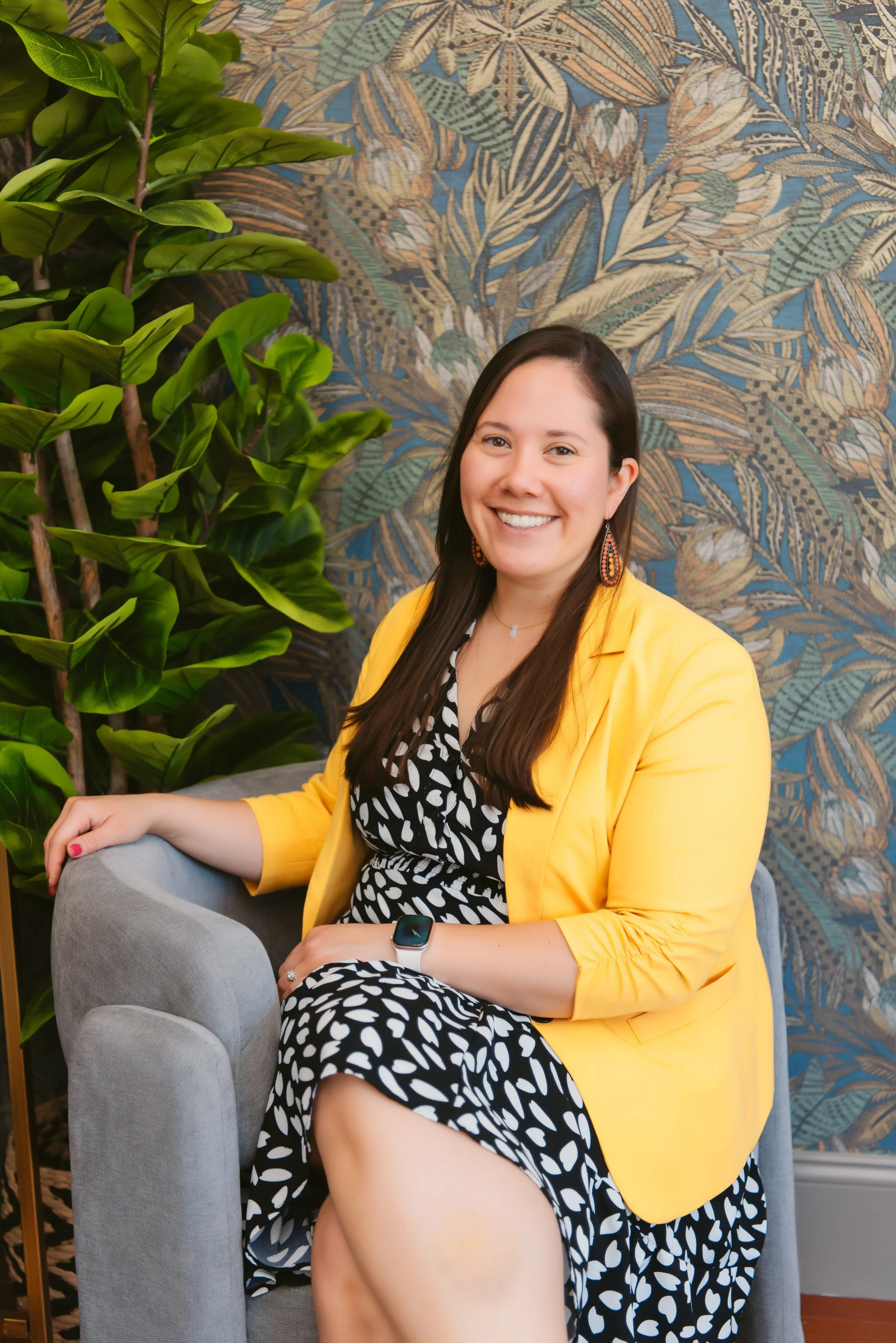 A smiling young woman with long dark hair, wearing a yellow blazer over a black and white polka dot dress, sitting on a light gray armchair next to a large leafy green plant, with a colorful leaf-patterned wallpaper in the background.