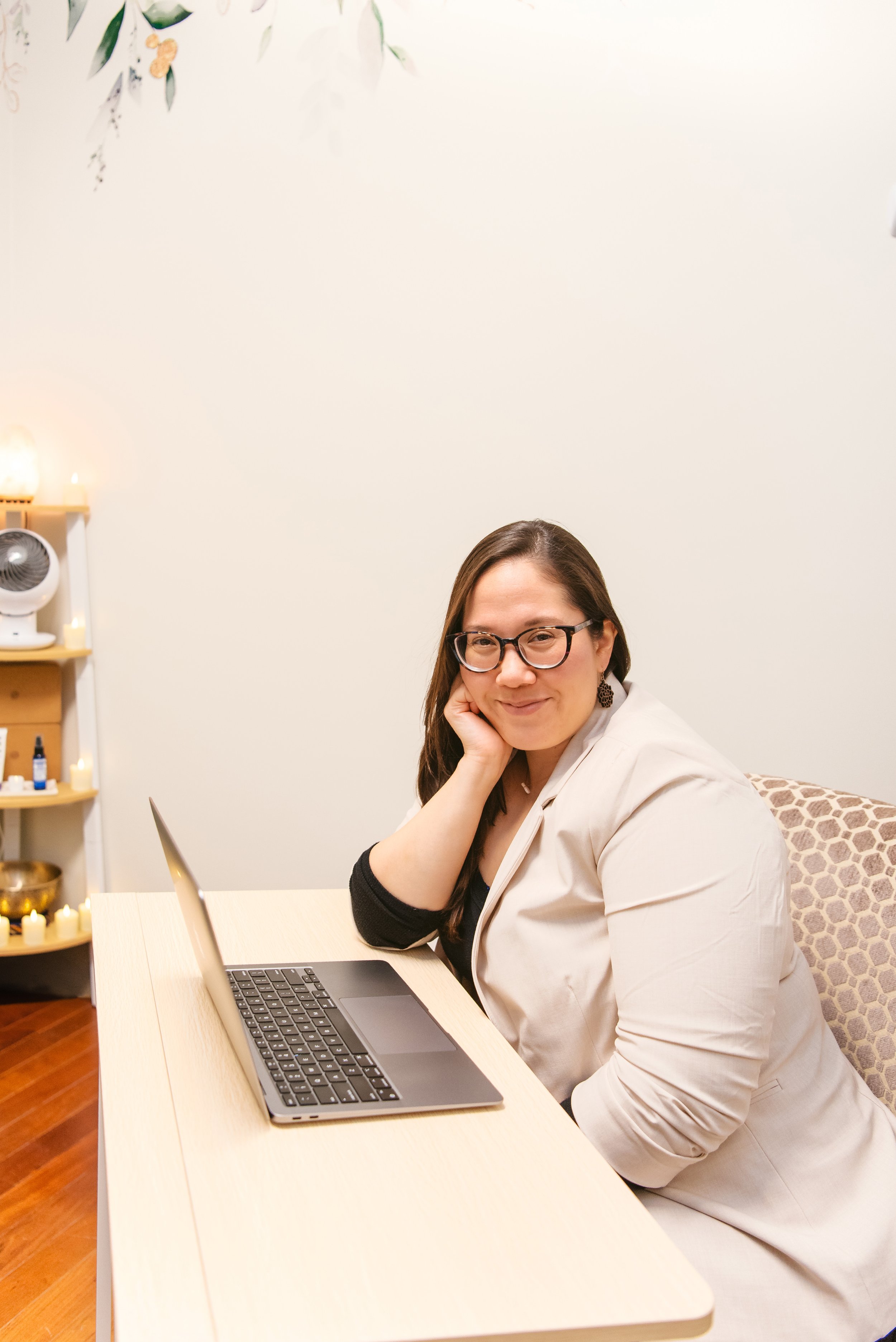 A woman with glasses and long brown hair sitting at a desk with a laptop, smiling and resting her head on her hand, in a room decorated with candles and shelves.