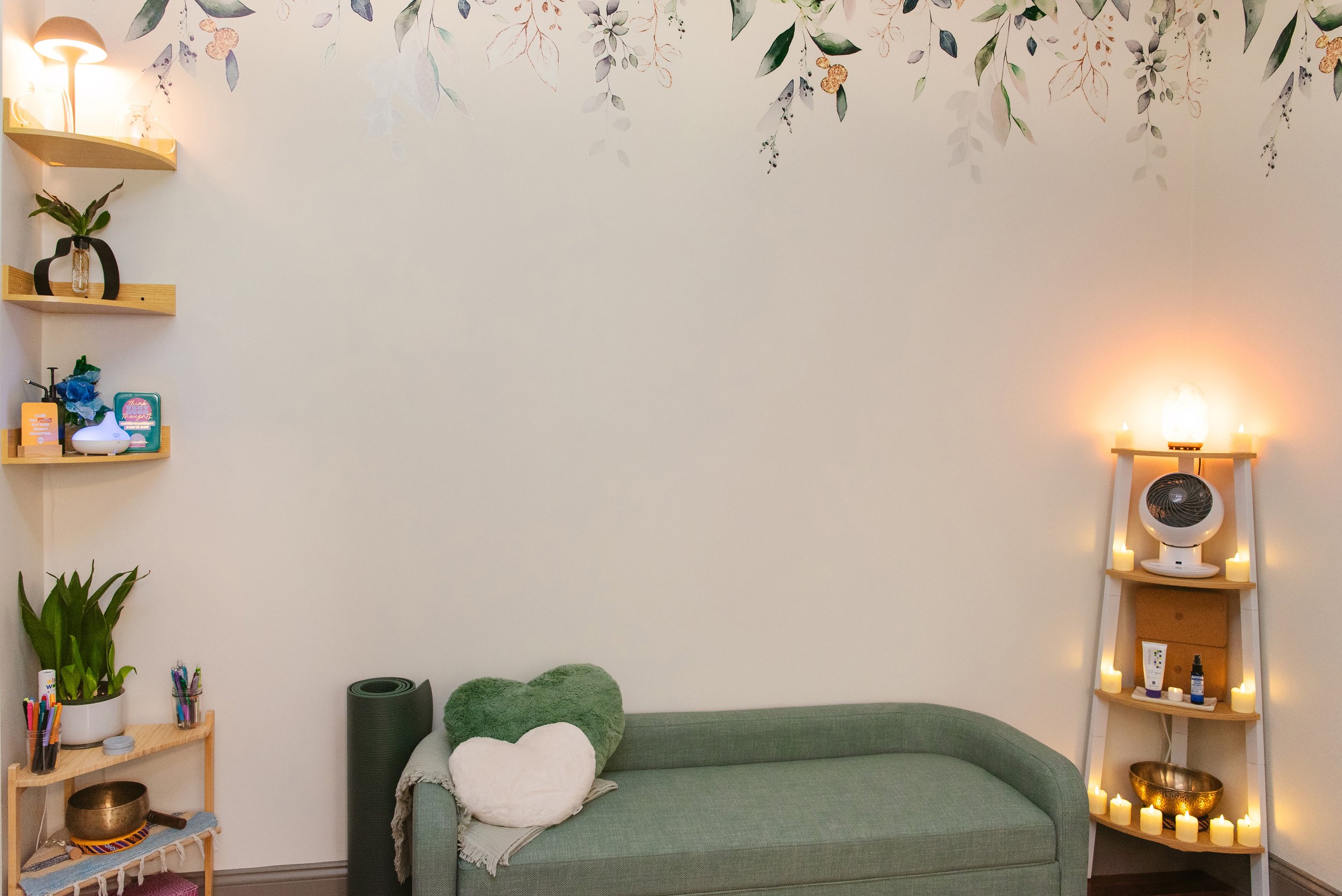 A cozy room with a light green sofa decorated with green and white heart-shaped pillows, a wall of white with a leafy floral border, a wooden shelf with plants and small decorative items, a side table with candles and candles, and a warm lamp.